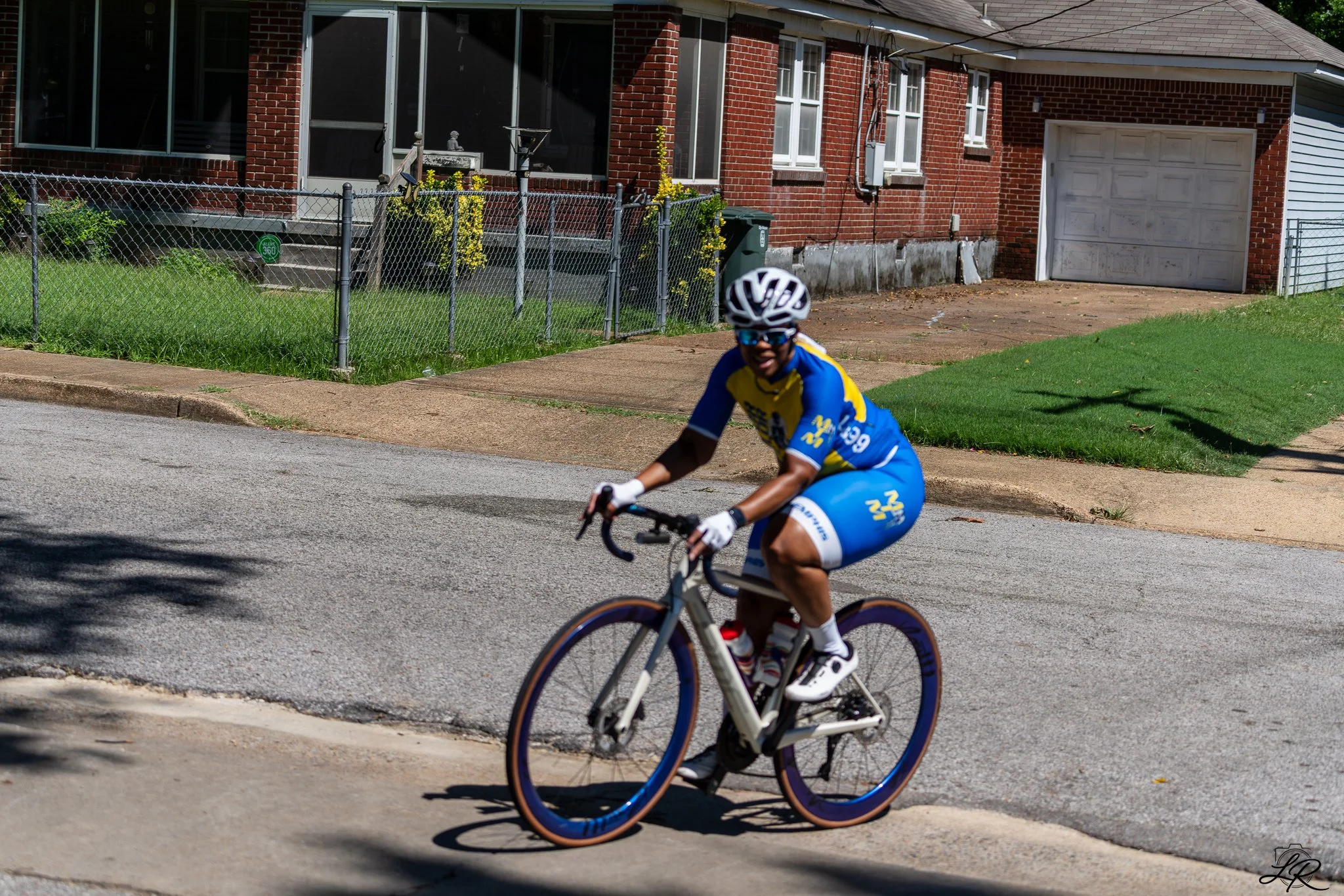 A person cycling on a residential street in front of a brick house with a garage, wearing a helmet and sunglasses, dressed in blue and yellow cycling gear.