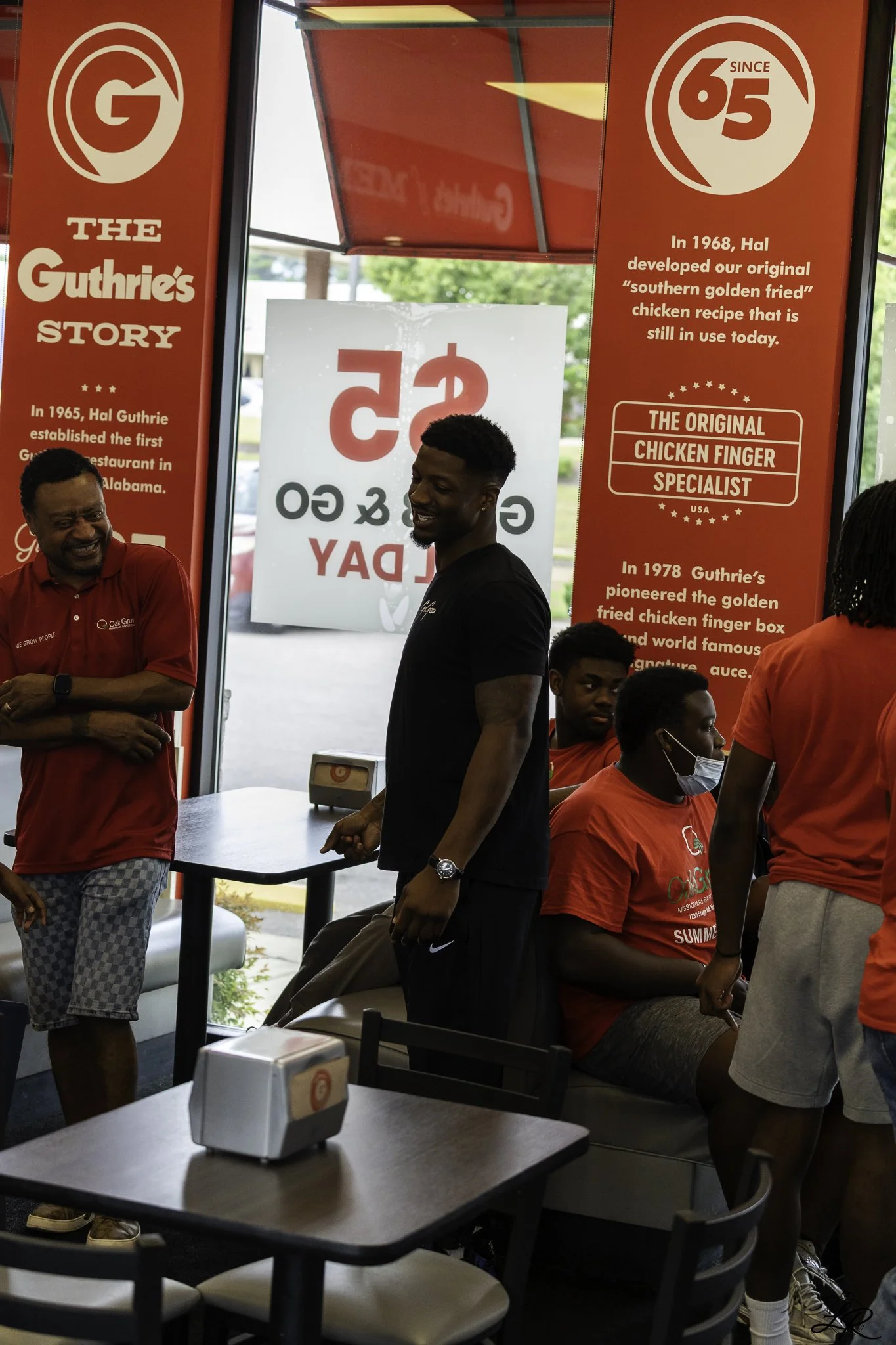 People inside a Guthrie's restaurant, a fast food chicken chain, with red banners displaying the history of the brand. Several individuals are talking and sitting, some wearing red shirts. The environment is casual with tables and chairs.