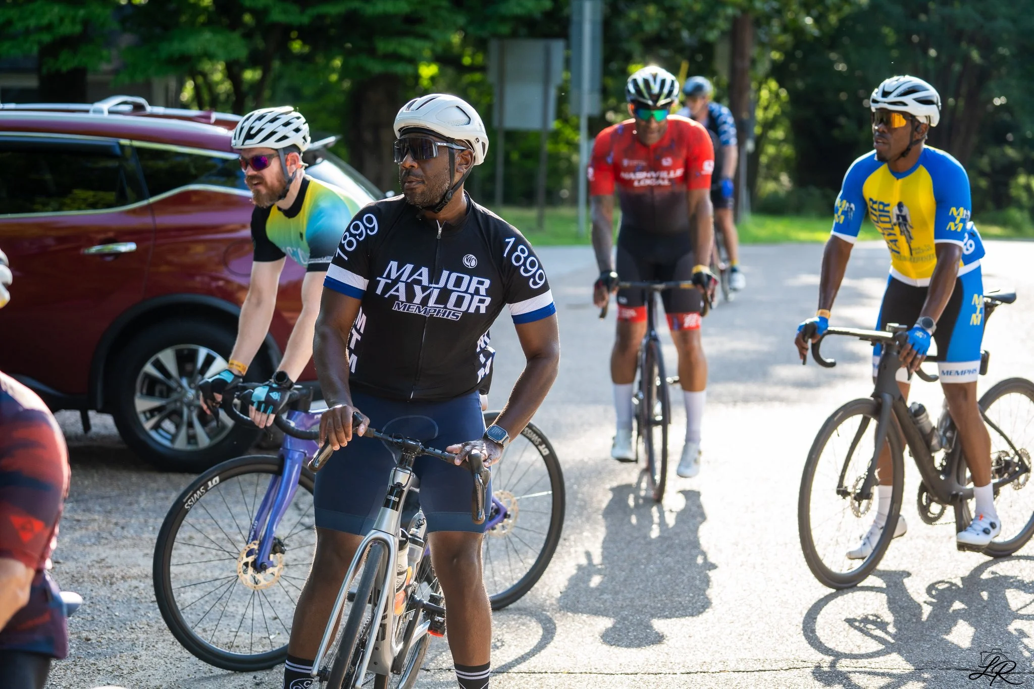 Group of cyclists in colorful jerseys and helmets riding on a sunny street with trees in the background.