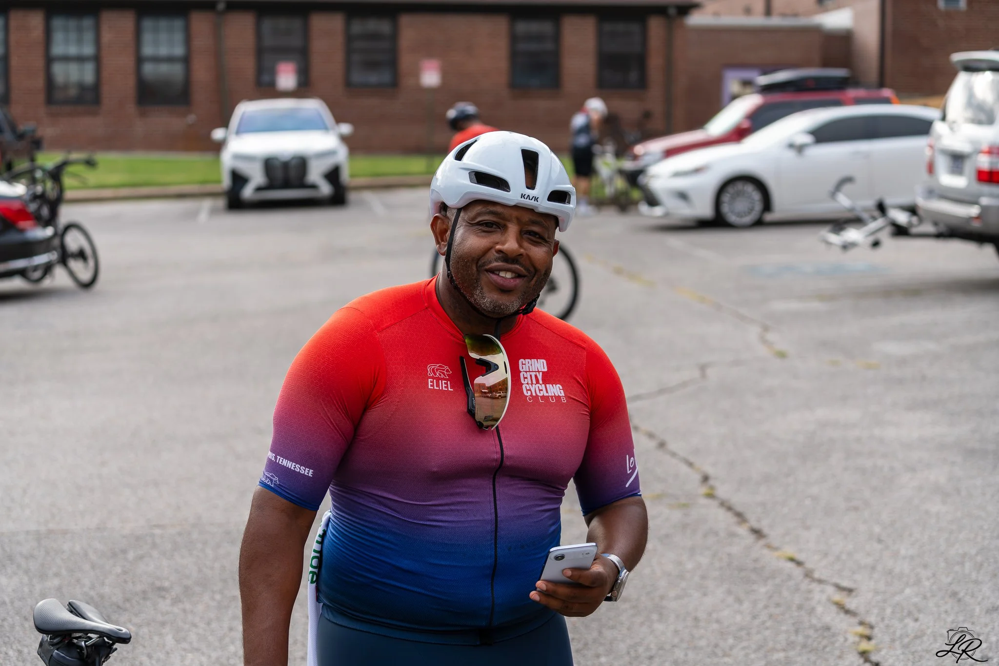 A man wearing a white bicycle helmet and a colorful cycling jersey holding a smartphone and standing next to a bicycle in a parking lot.