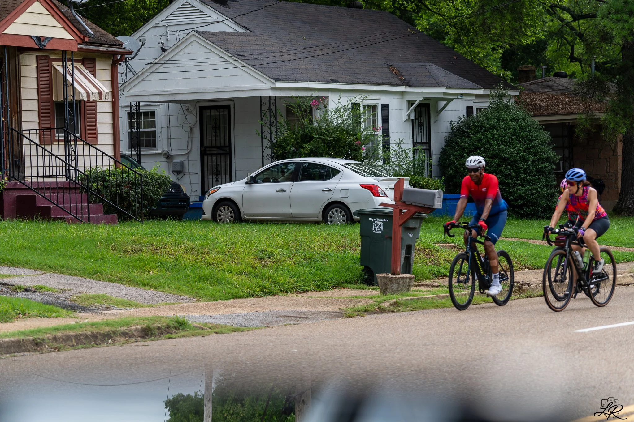 Two people riding bicycles on a residential sidewalk, with houses, cars, bushes, and trees in the background.