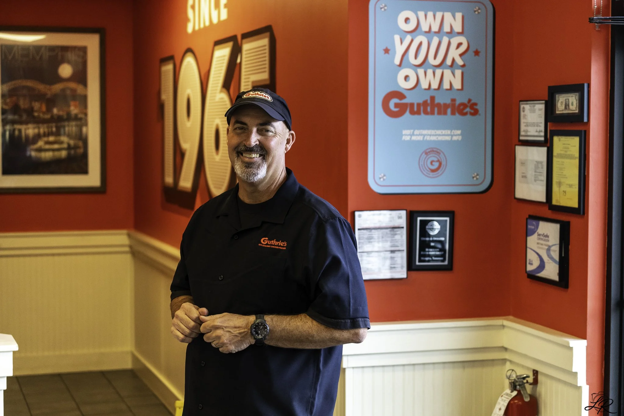 A smiling man in a black Guthrie's uniform and cap standing inside a Guthrie's restaurant, with red walls decorated with framed pictures and signs, including a blue sign saying "Own Your Own Guthrie's."