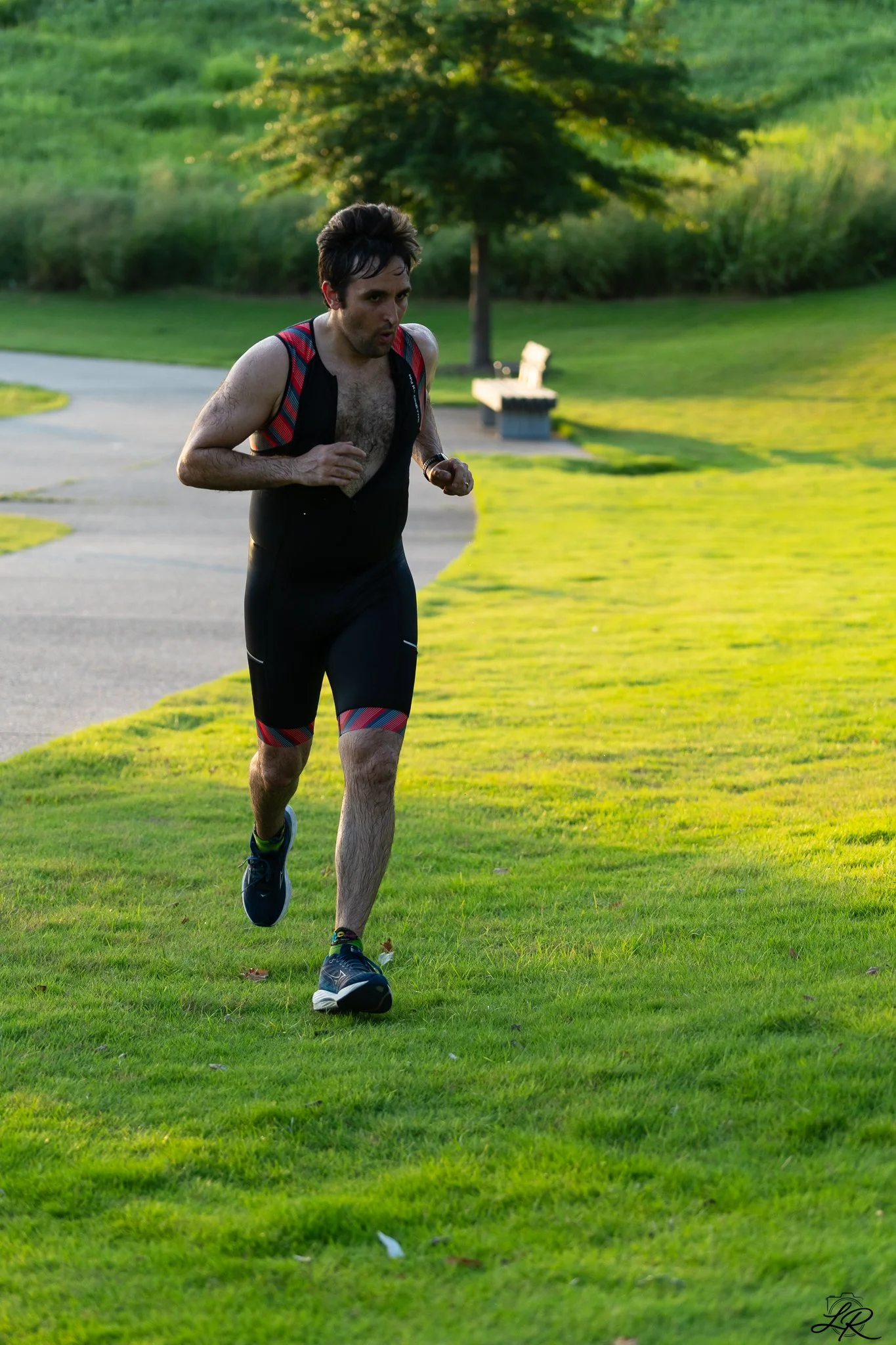 A man running outdoors on a grassy path with trees and shrubs in the background, wearing a sleeveless athletic shirt and shorts, with a determined expression.