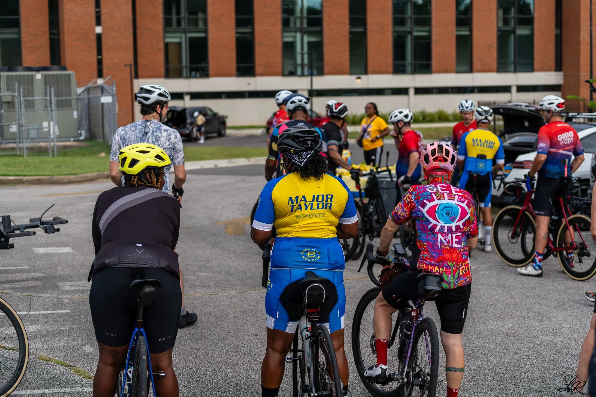 A group of cyclists gathered outdoors in front of a modern brick building, many wearing helmets and colorful cycling jerseys, preparing for a ride.