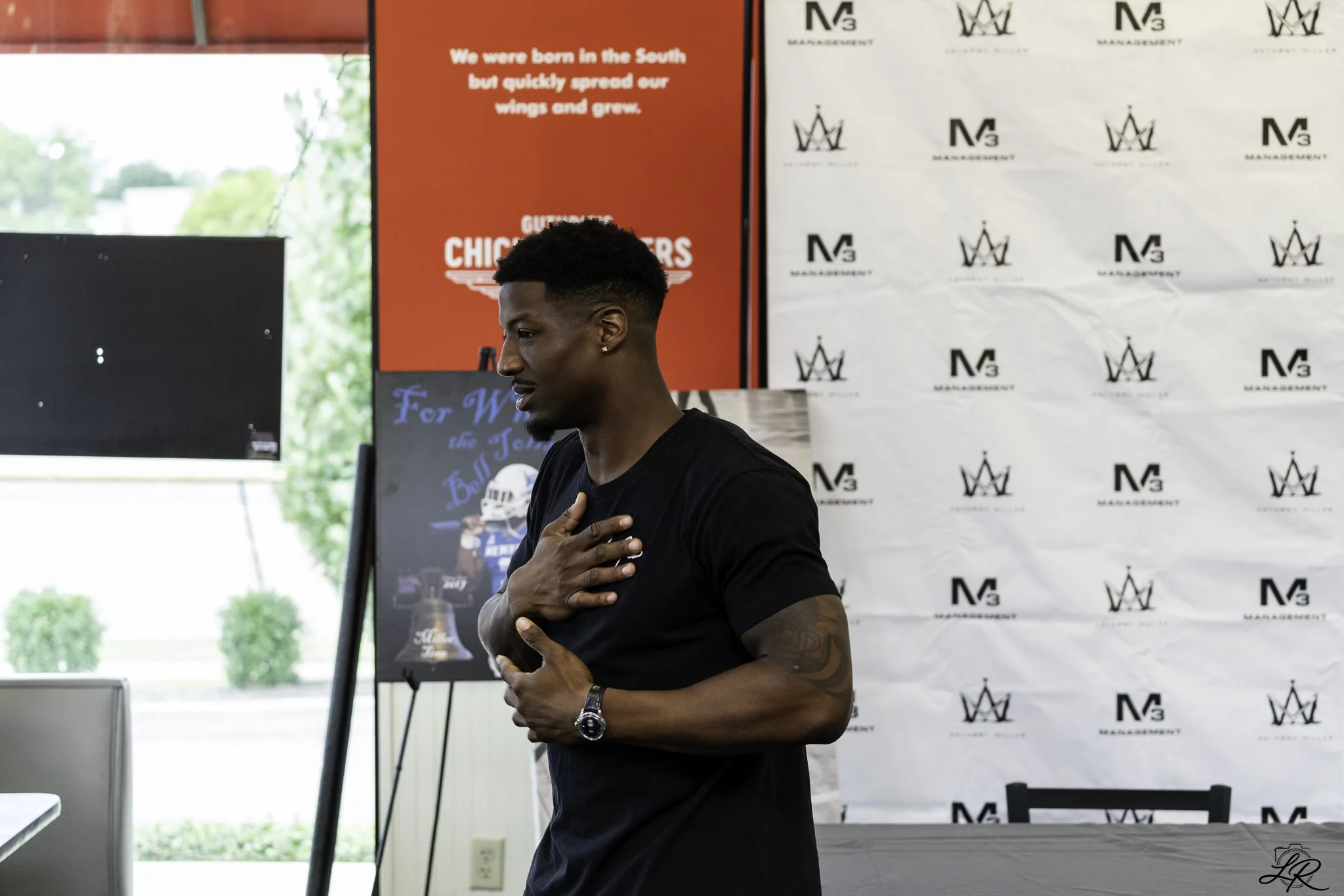 A young man with short black hair and a tattoo on his arm stands with his hand over his heart in an indoor setting with banners in the background, one red with white text and another white with black logos.
