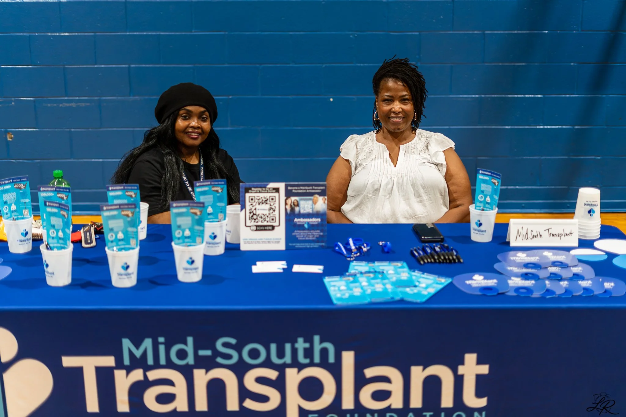 Two women seated at a table displaying promotional materials for the Mid-South Transplant Foundation, with a blue wall in the background.
