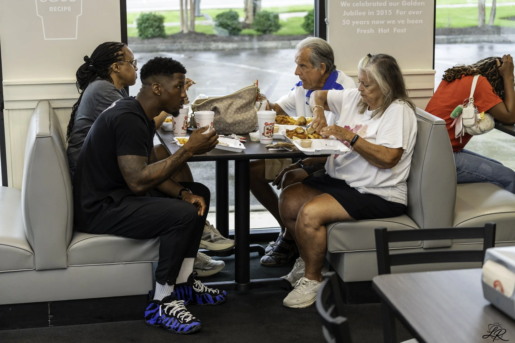 People sitting inside a fast-food restaurant, eating lunch and talking, with large windows showing an outdoor parking lot and green landscape.