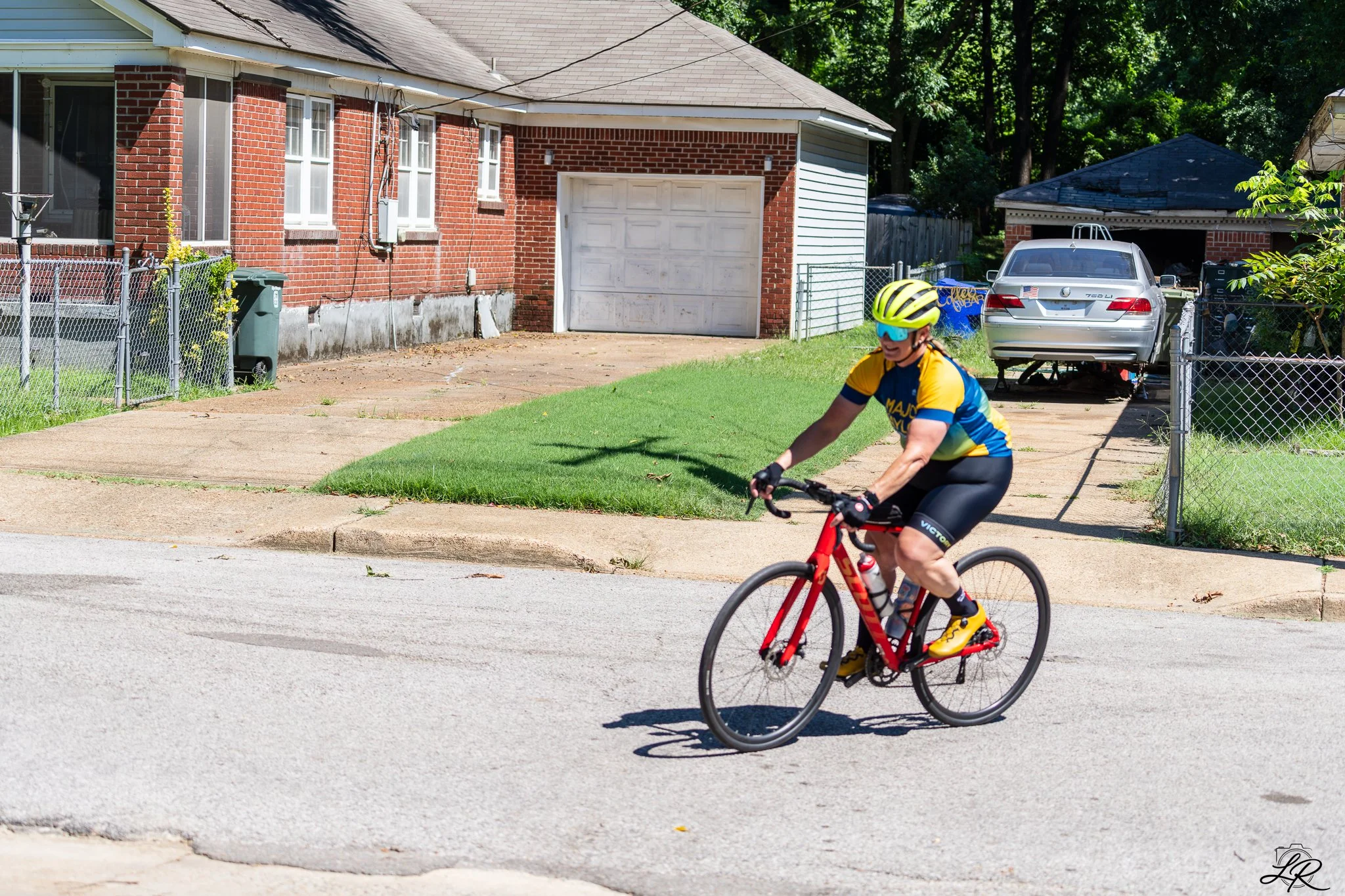 A boy riding a red bicycle on a residential street, wearing a yellow and blue cycling helmet, sunglasses, and a blue and yellow cycling kit. A brick house with a white garage door, trees, and a parked car are visible in the background.