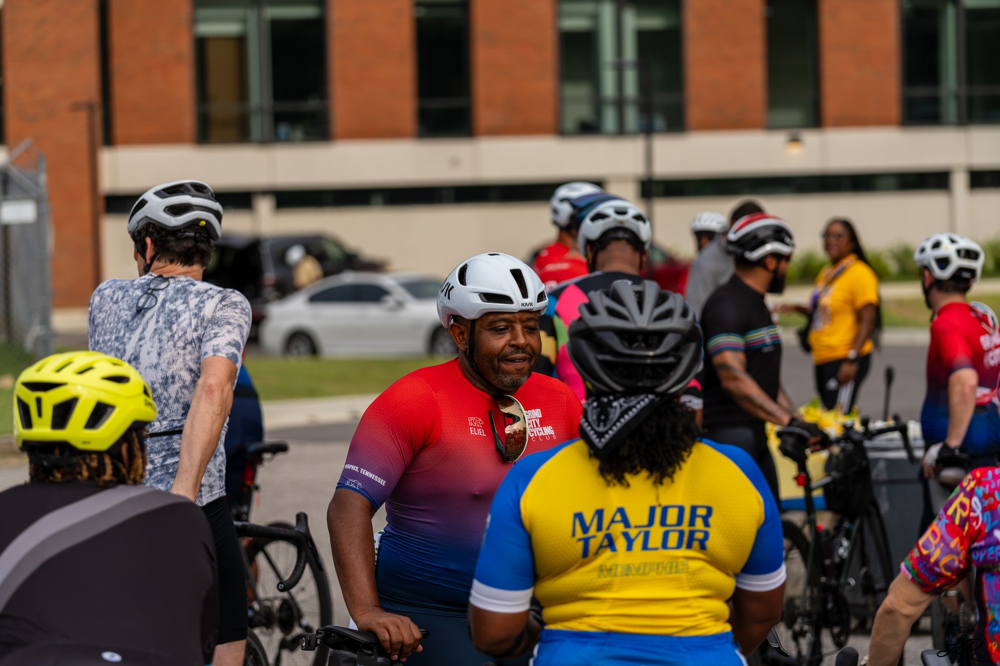 A group of cyclists, some wearing helmets, gathered outdoors in front of a modern building with brick and glass exterior. The man in the foreground is wearing a red and purple cycling jersey with "Major Taylor" written on the back and a white helmet.