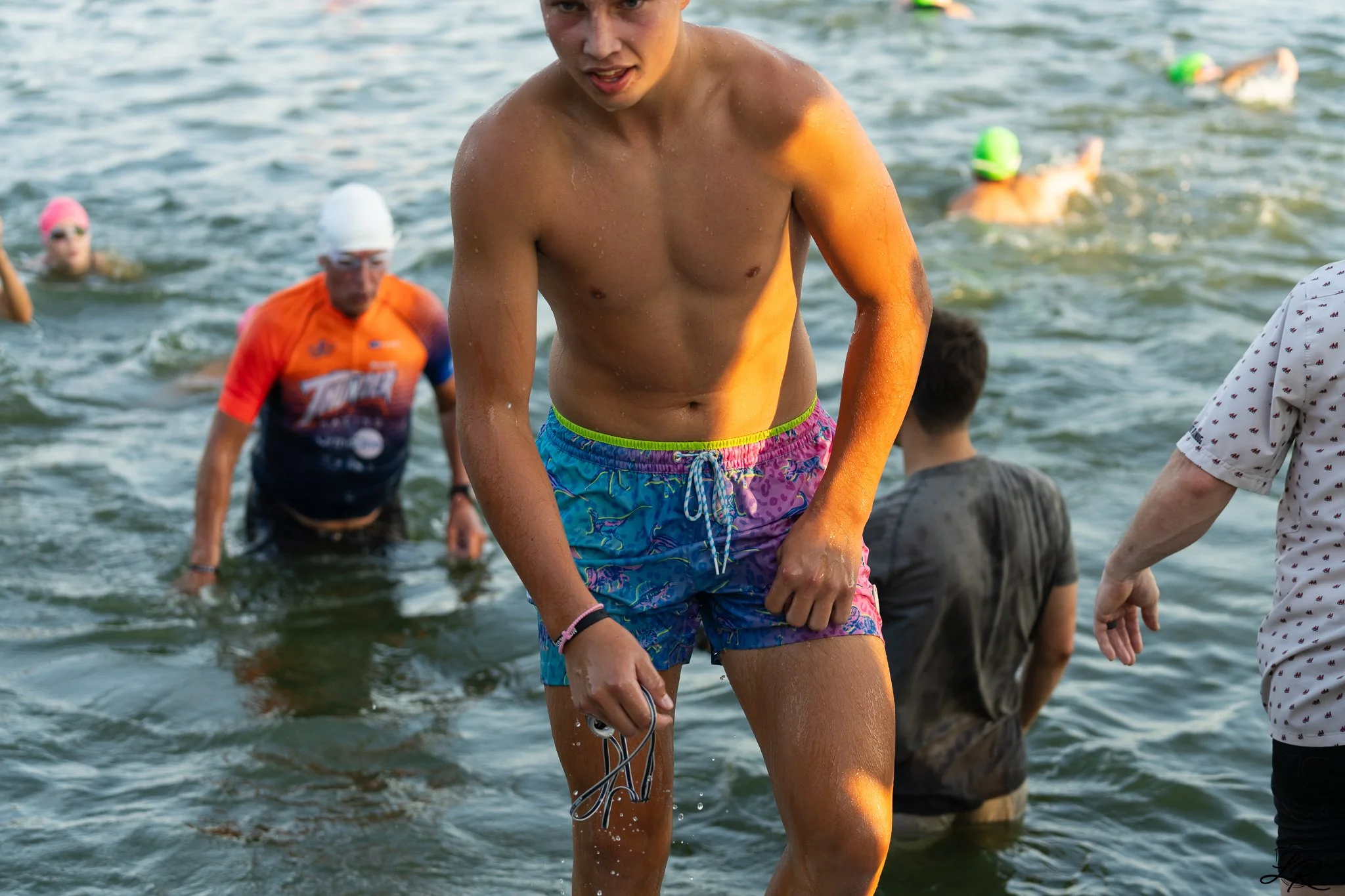 Young man in colorful swim trunks emerging from a body of water during a swimming event, with other swimmers and people in the background.
