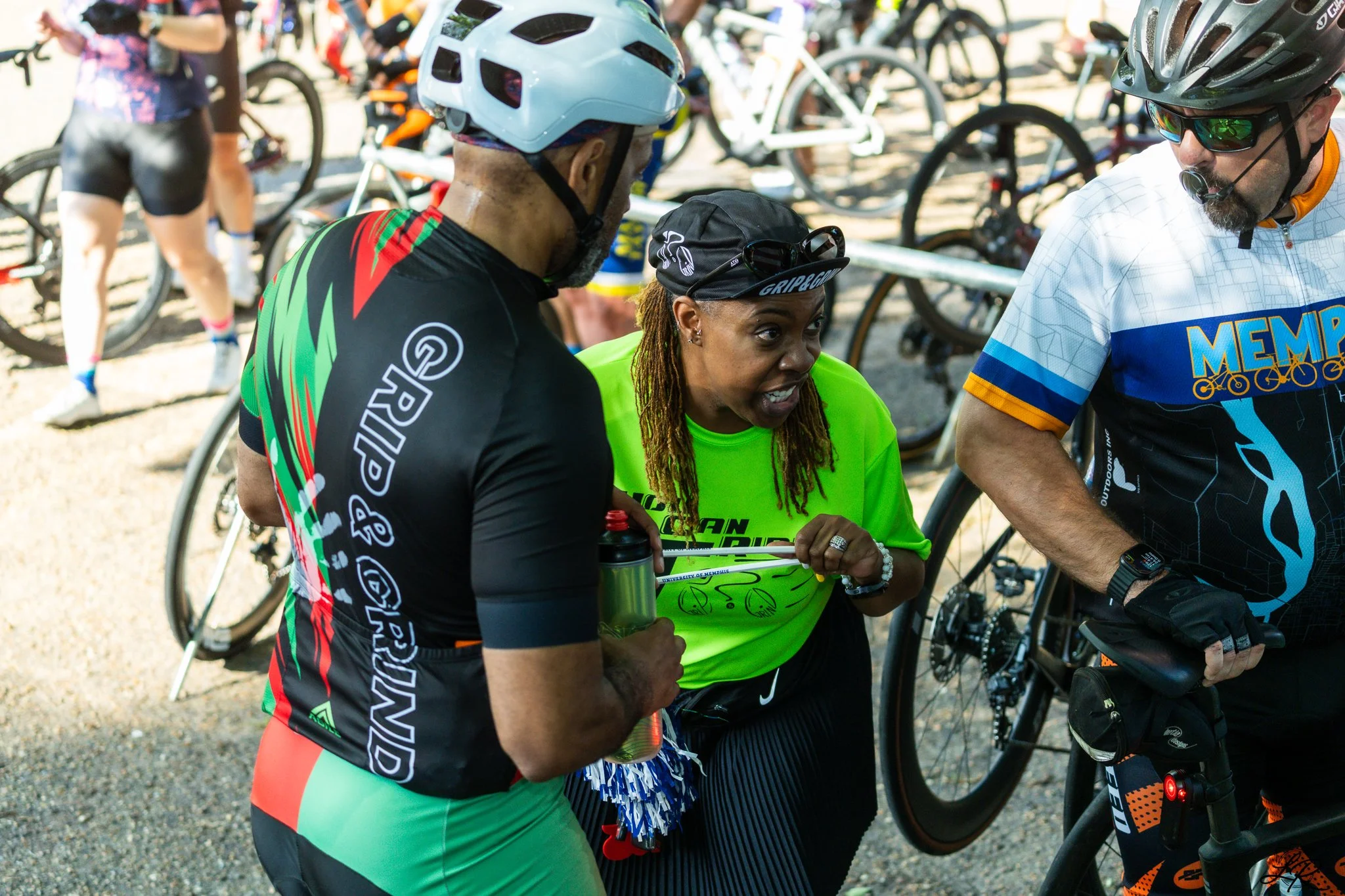 A group of cyclists at a biking event, with one woman talking to two men, surrounded by bikes and other participants.