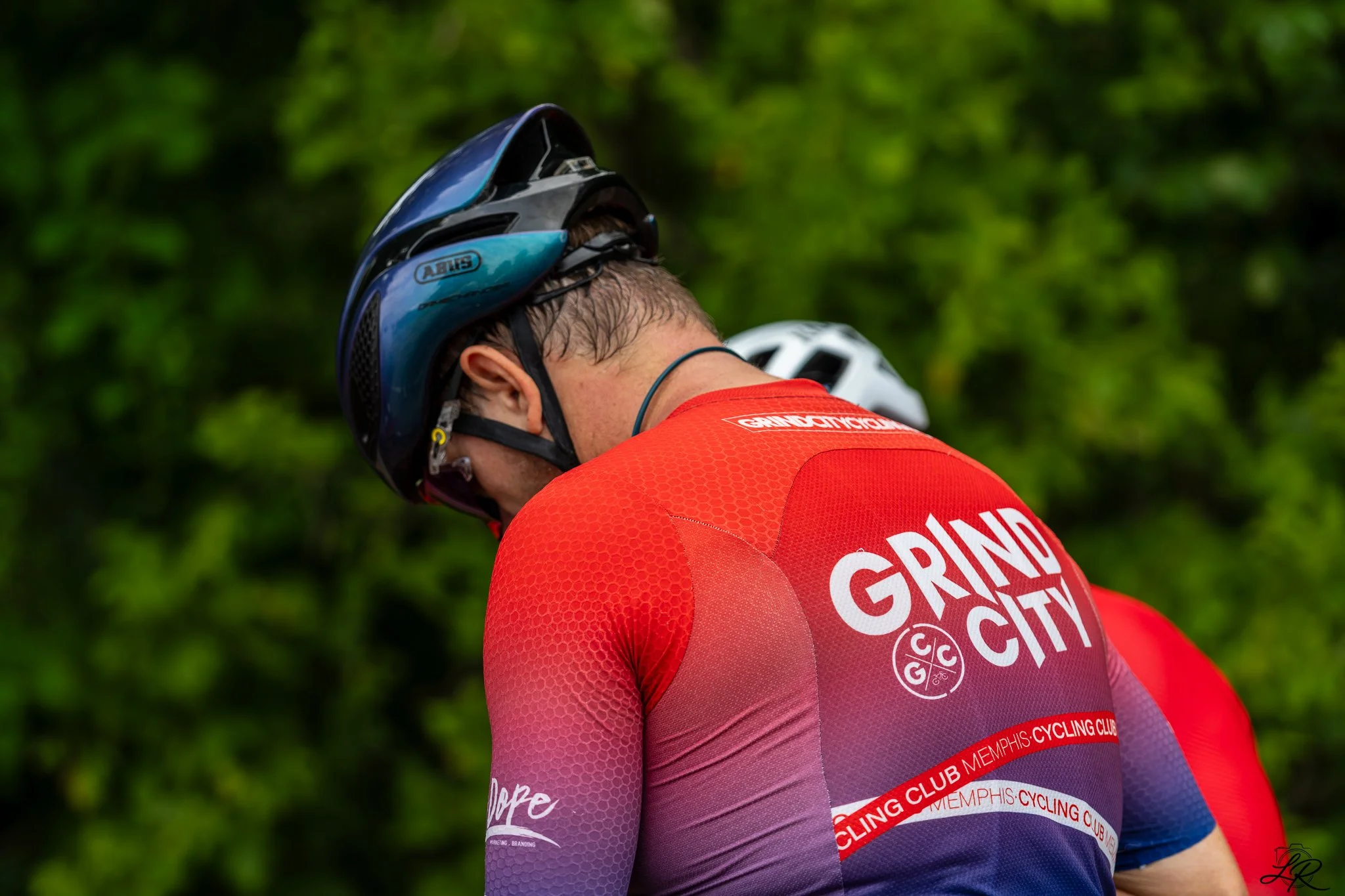 A cyclist in a red and purple Memphis Cycling Club jersey and a black and blue helmet, looking downward, outdoors with green trees in the background.