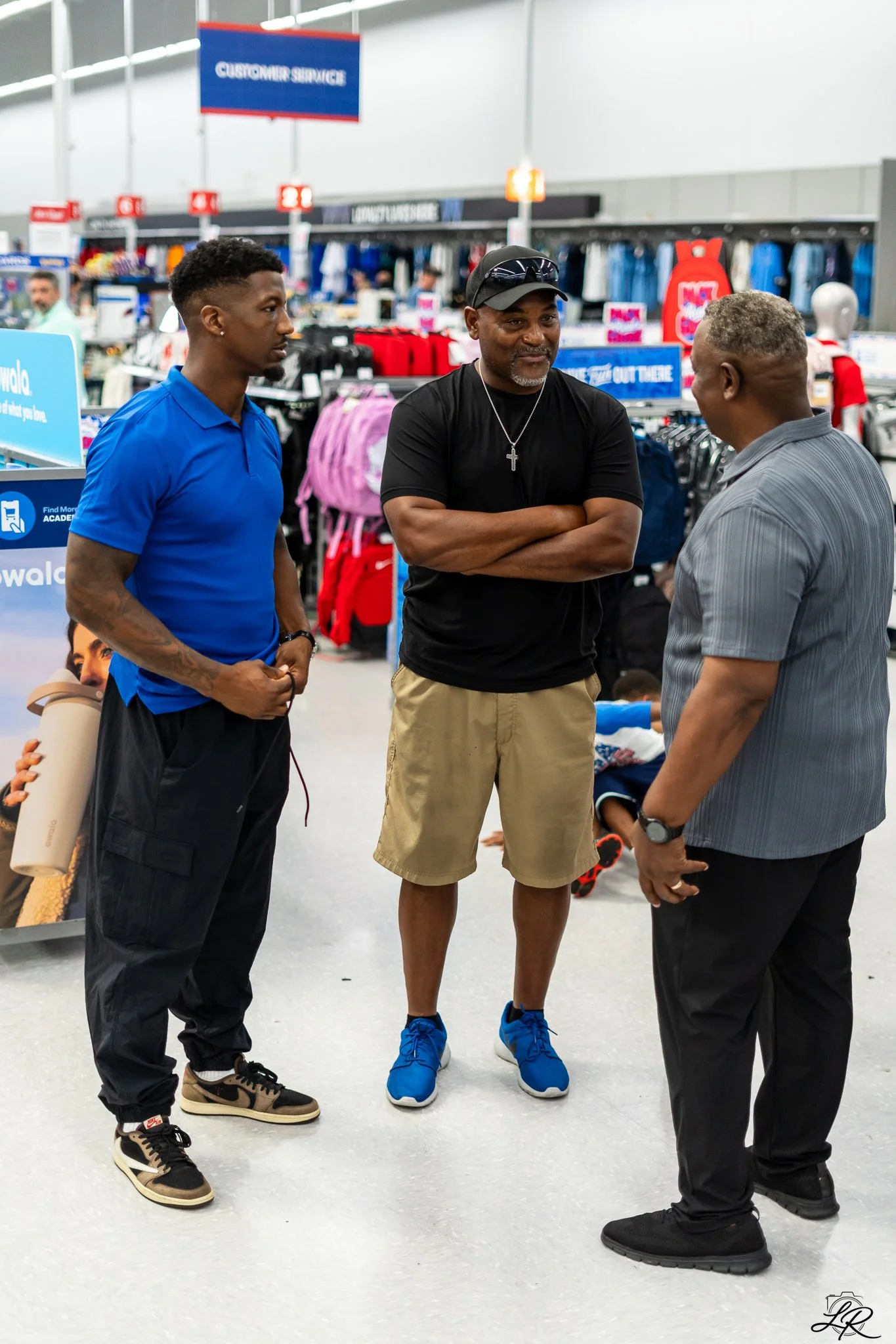 Three men stand together in a retail store, engaged in conversation. One wears a blue polo shirt, the second wears a black shirt with a cross necklace and tan shorts, and the third wears a gray shirt with black pants. The store has clothing and acces