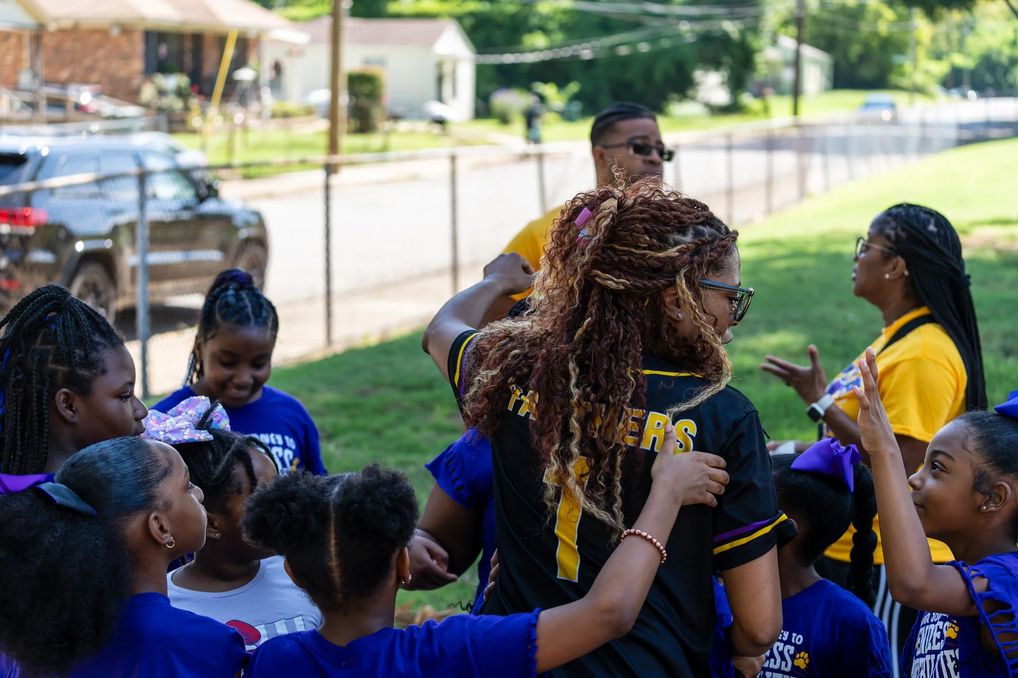 A group of children in blue uniforms standing outdoors on a sunny day, some with ribbons in their hair, gathered around a woman with glasses and curly hair. The woman is embracing or greeting a girl, and others are smiling and watching. There are hou