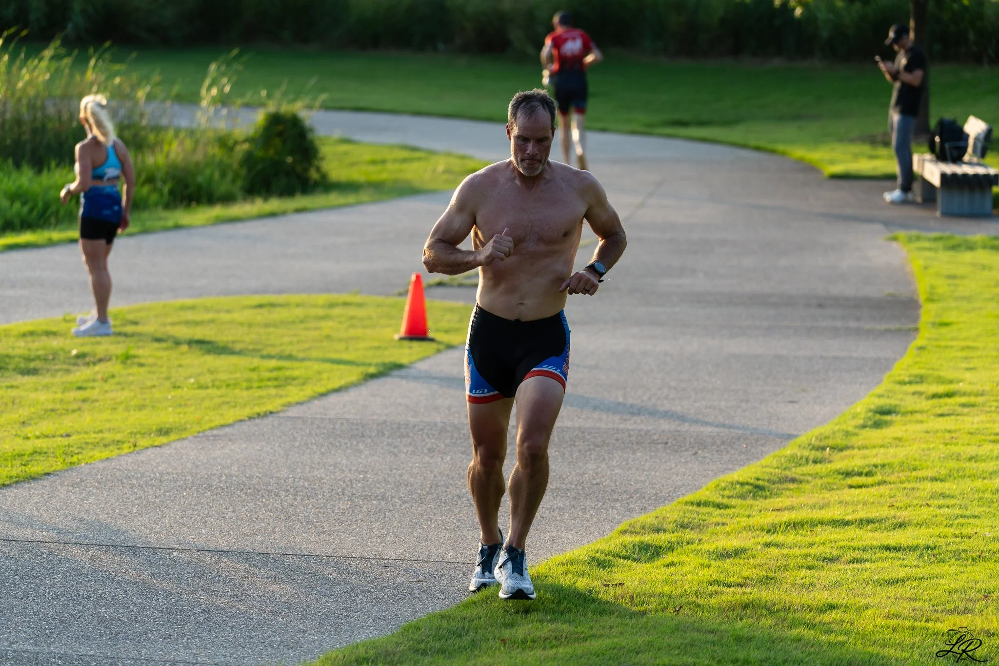A shirtless man running outdoors on a paved path with some people in the background, including a woman in athletic clothing and a person with a phone, during daylight.