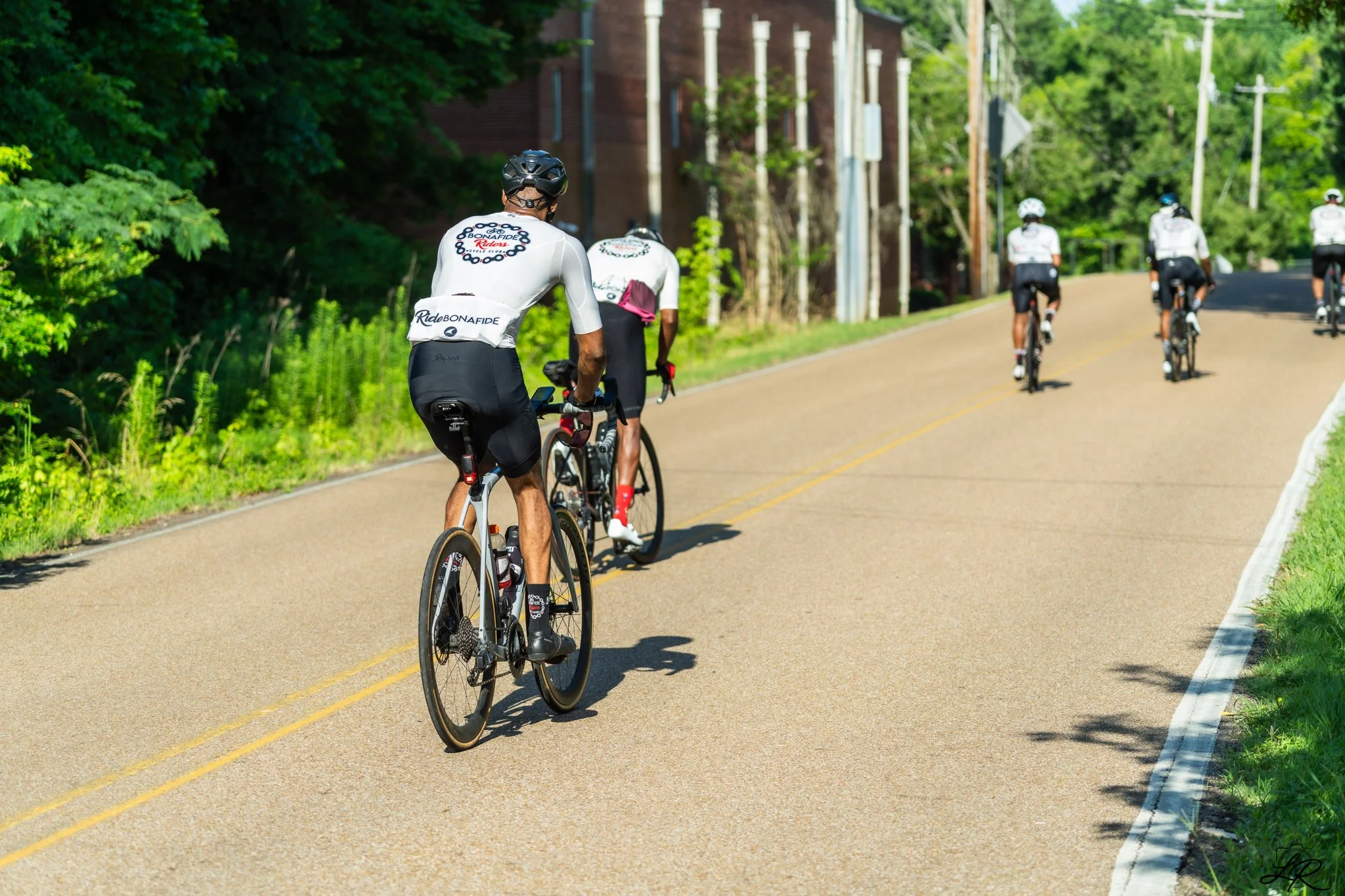 A group of cyclists riding on a paved road surrounded by green trees under a sunny sky.