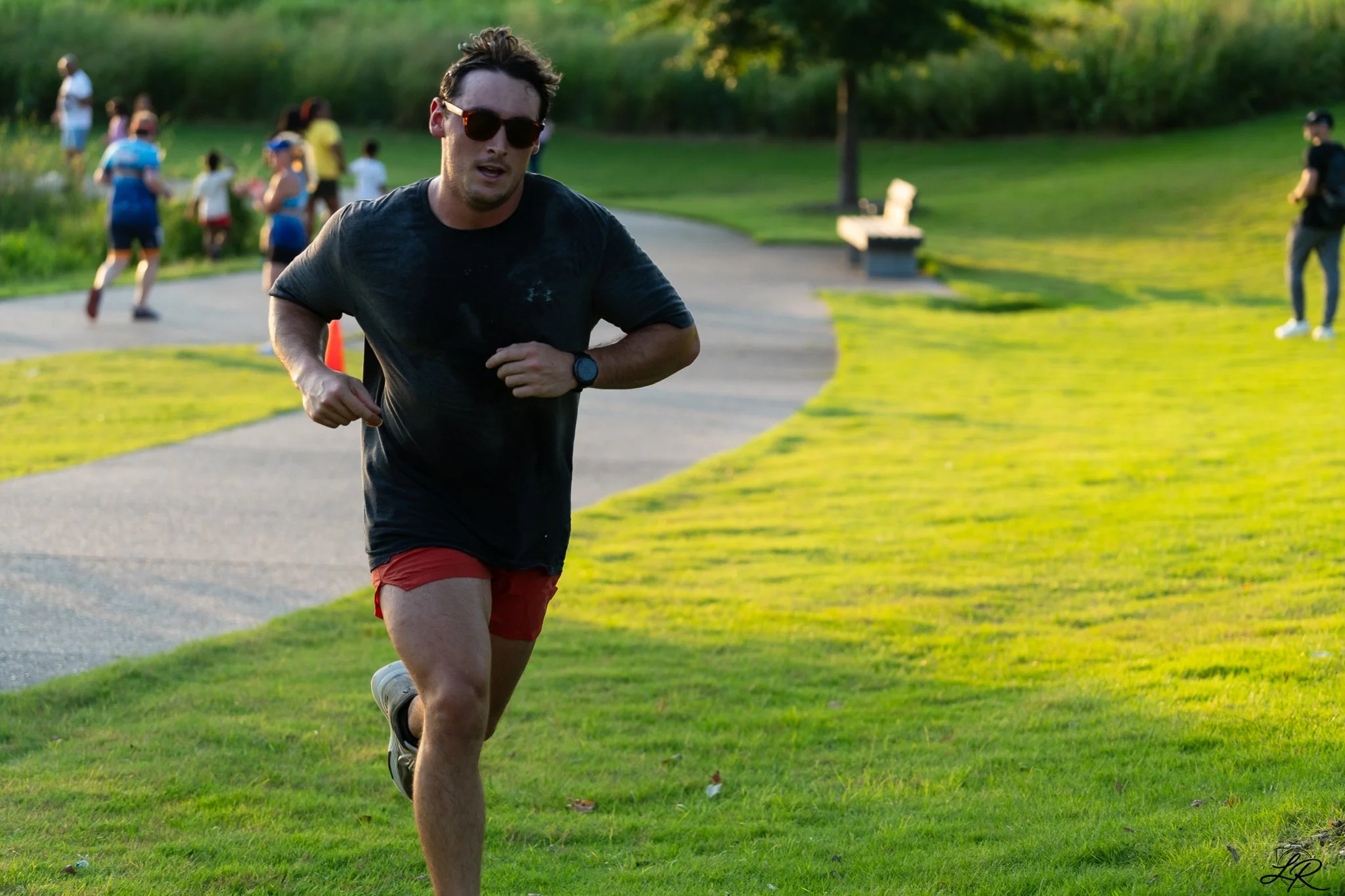 A man running in a park during daylight, wearing a black shirt, red shorts, sunglasses, and a watch, with several people in the background near trees and grass.