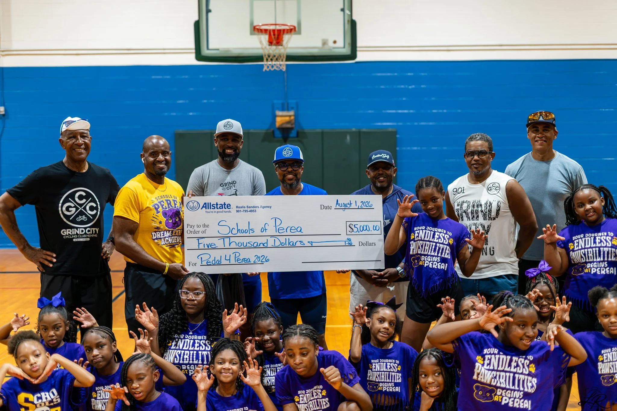 A group of children and adults in a gymnasium holding a large check for $5,000 made out to Schools of Perea, dated August 1, 2026, with the sender Realms Sanders Agency. The people are smiling, some wearing blue T-shirts with messages about endless p