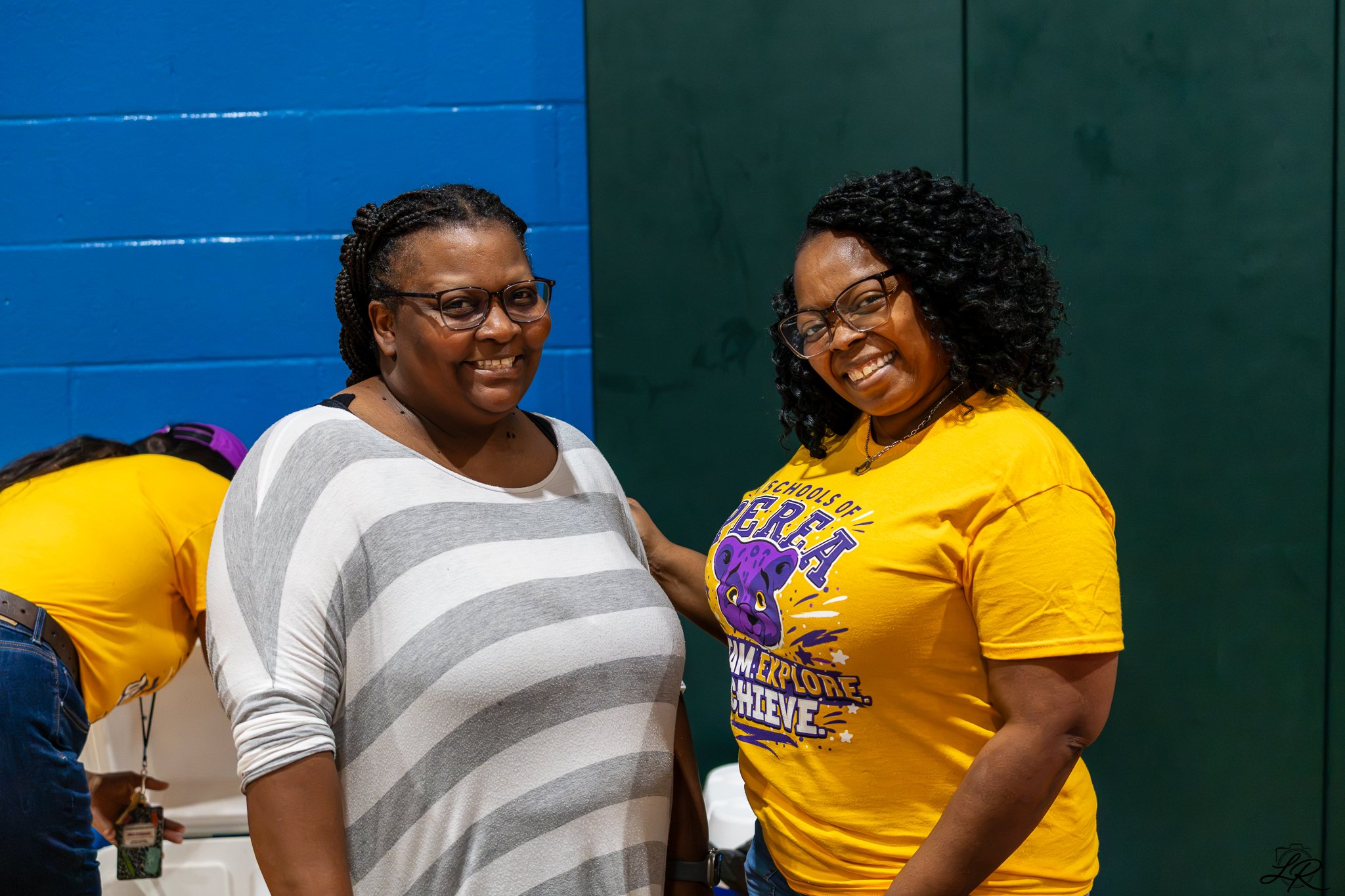 Two women with glasses smiling at each other, one wearing a gray and white striped shirt and the other wearing a yellow T-shirt with purple lettering and a graphic, in an indoor gymnasium setting.