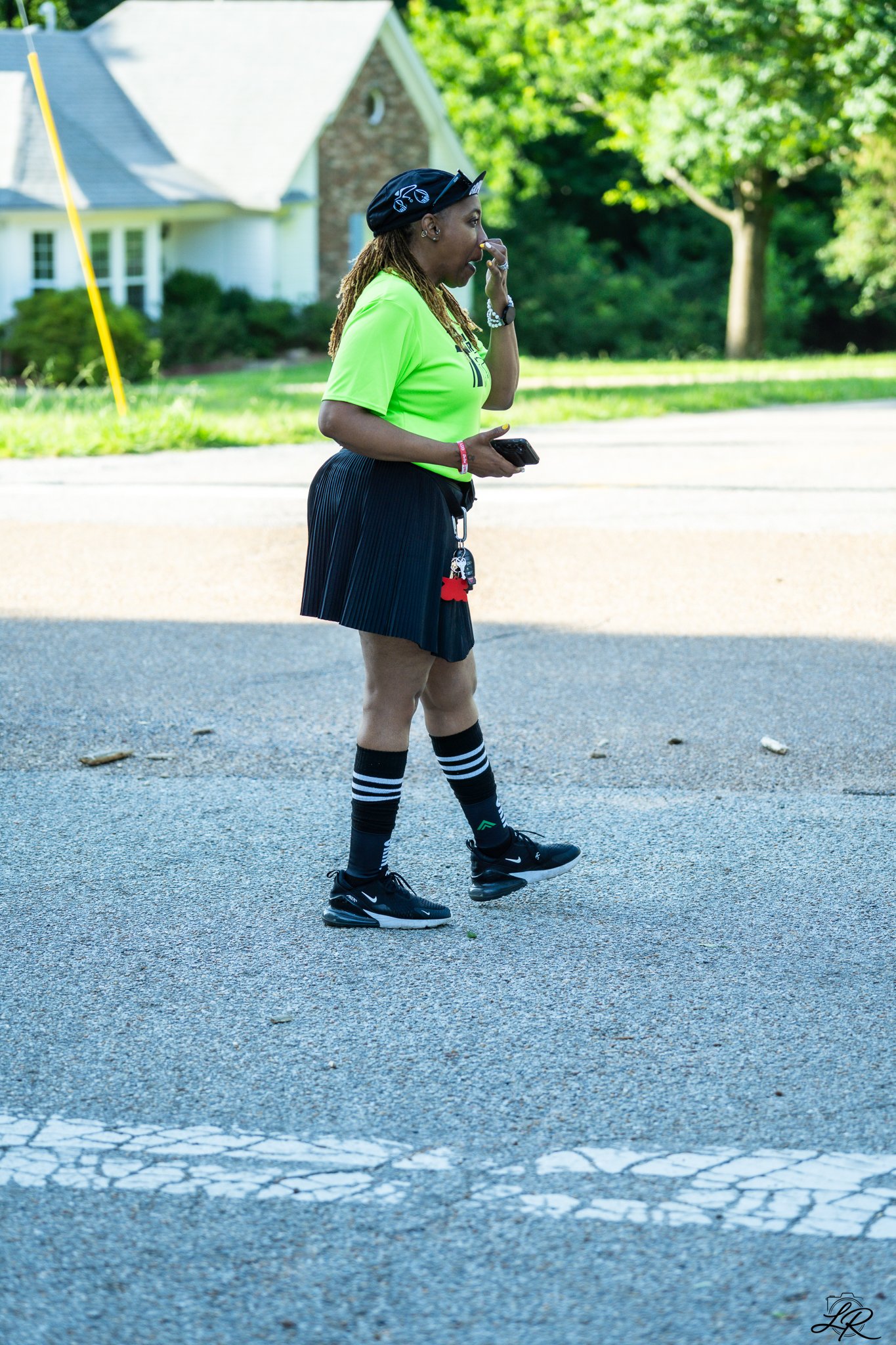 A woman dressed in a bright yellow shirt, black pleated shorts, knee-high striped socks, and black sneakers stands on a street with trees and a house in the background. She is holding a phone in her left hand and touching her nose with her right hand