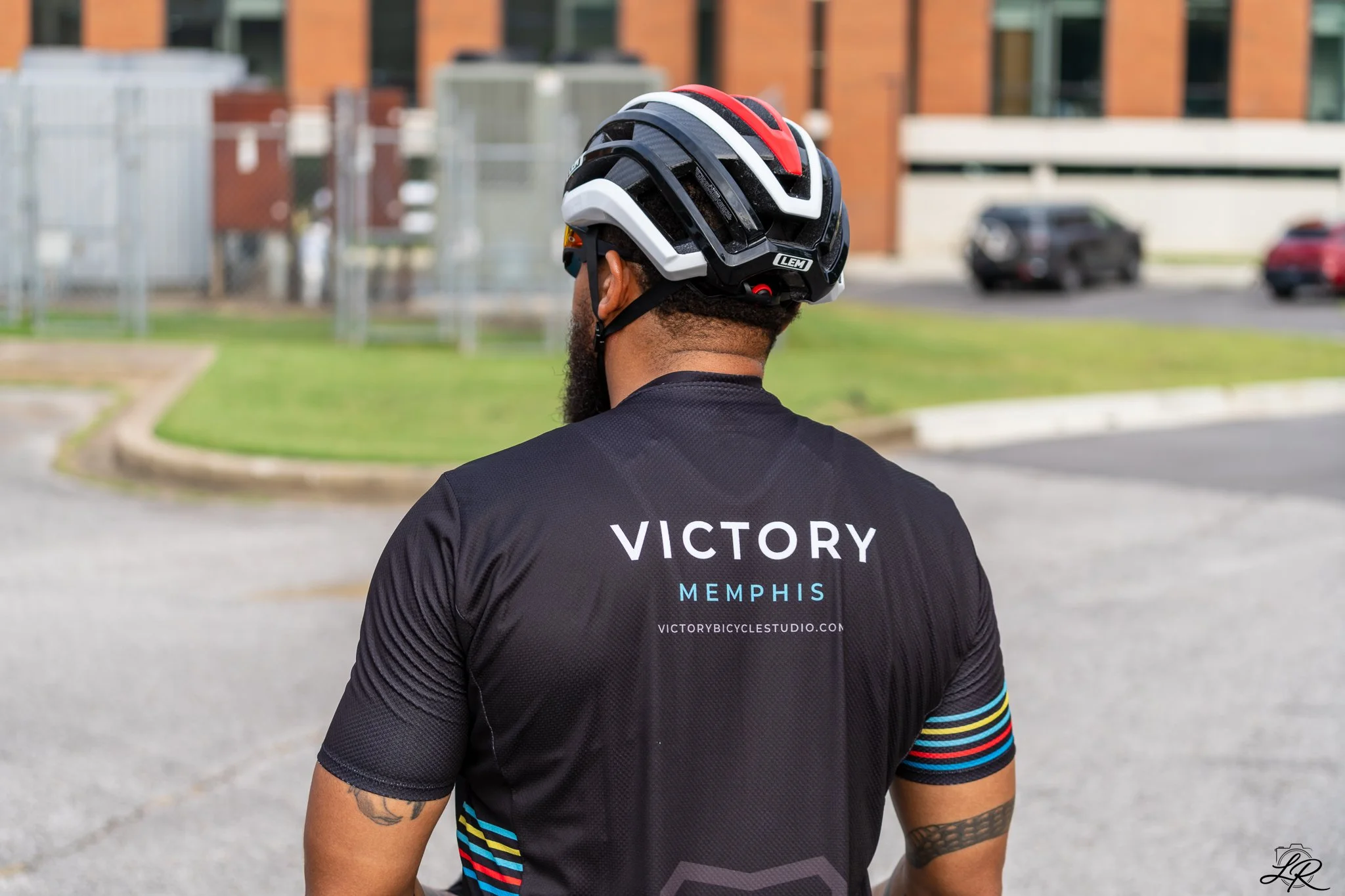 Man wearing a black cycling jersey with "Victory Memphis" logo and a black, white, and red helmet, standing outdoors with parking lot and buildings in the background.