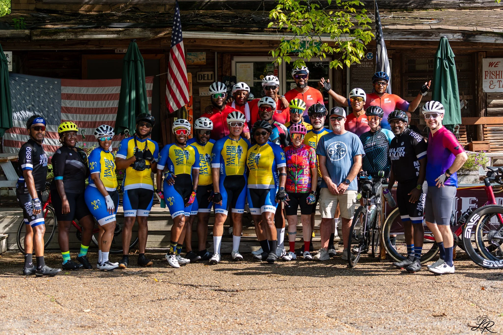A group of cyclists wearing colorful jerseys and helmets standing together outside a wooden building with American flags and green umbrellas. They are smiling and posing for the photo, with some holding their bikes.