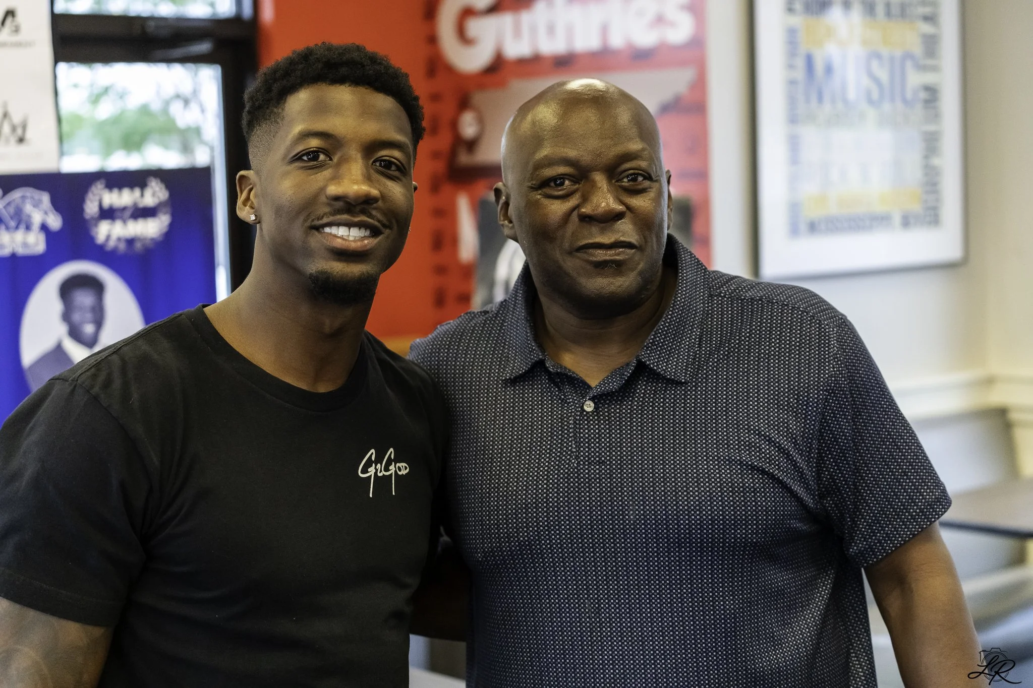 Two men standing close together indoors, smiling at the camera. One man has dark skin, is bald, and wears a dark patterned shirt. The other man has dark skin, short curly hair, a goatee, and is wearing a black t-shirt with white writing. There are si