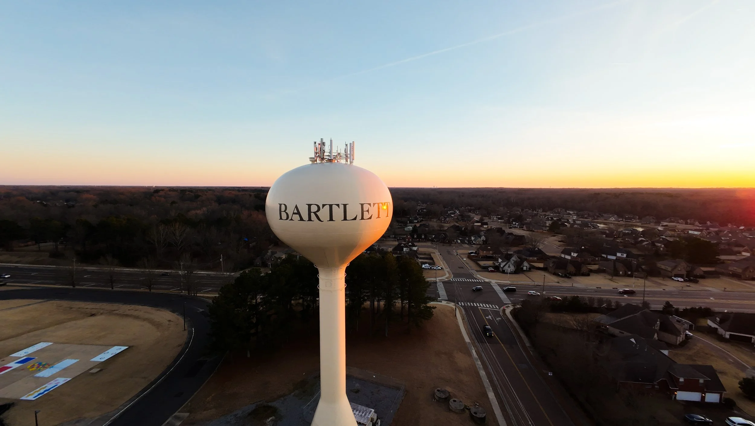 A white water tower with the words 'BARTLETT' painted on it, situated above a suburban area with houses and roads during sunset.