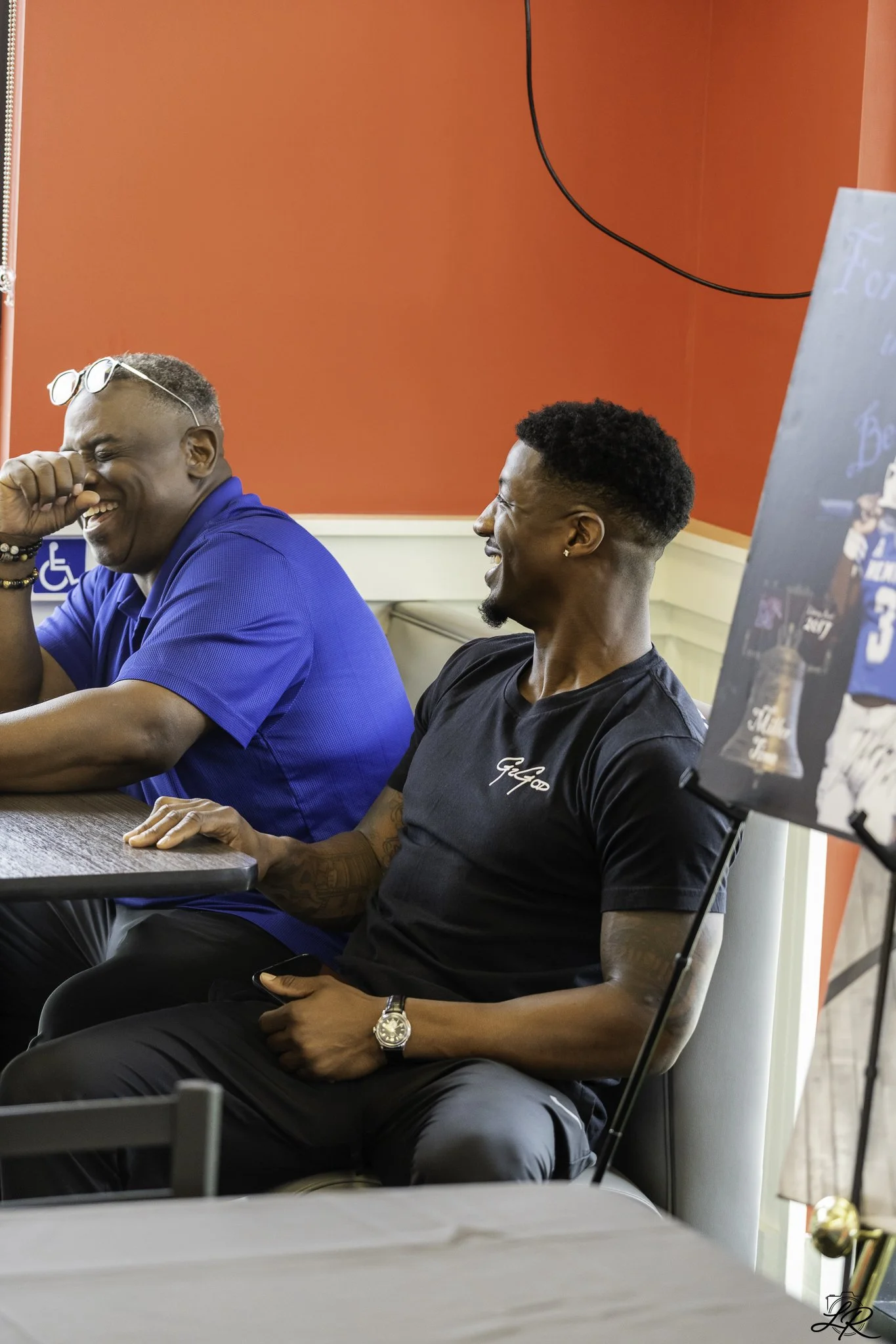 Two men seated at a table in a room with red walls, laughing and having a friendly conversation. One man is wearing a blue shirt and sunglasses on his head, while the other is wearing a black T-shirt and a wristwatch. A sign with a wheelchair symbol 