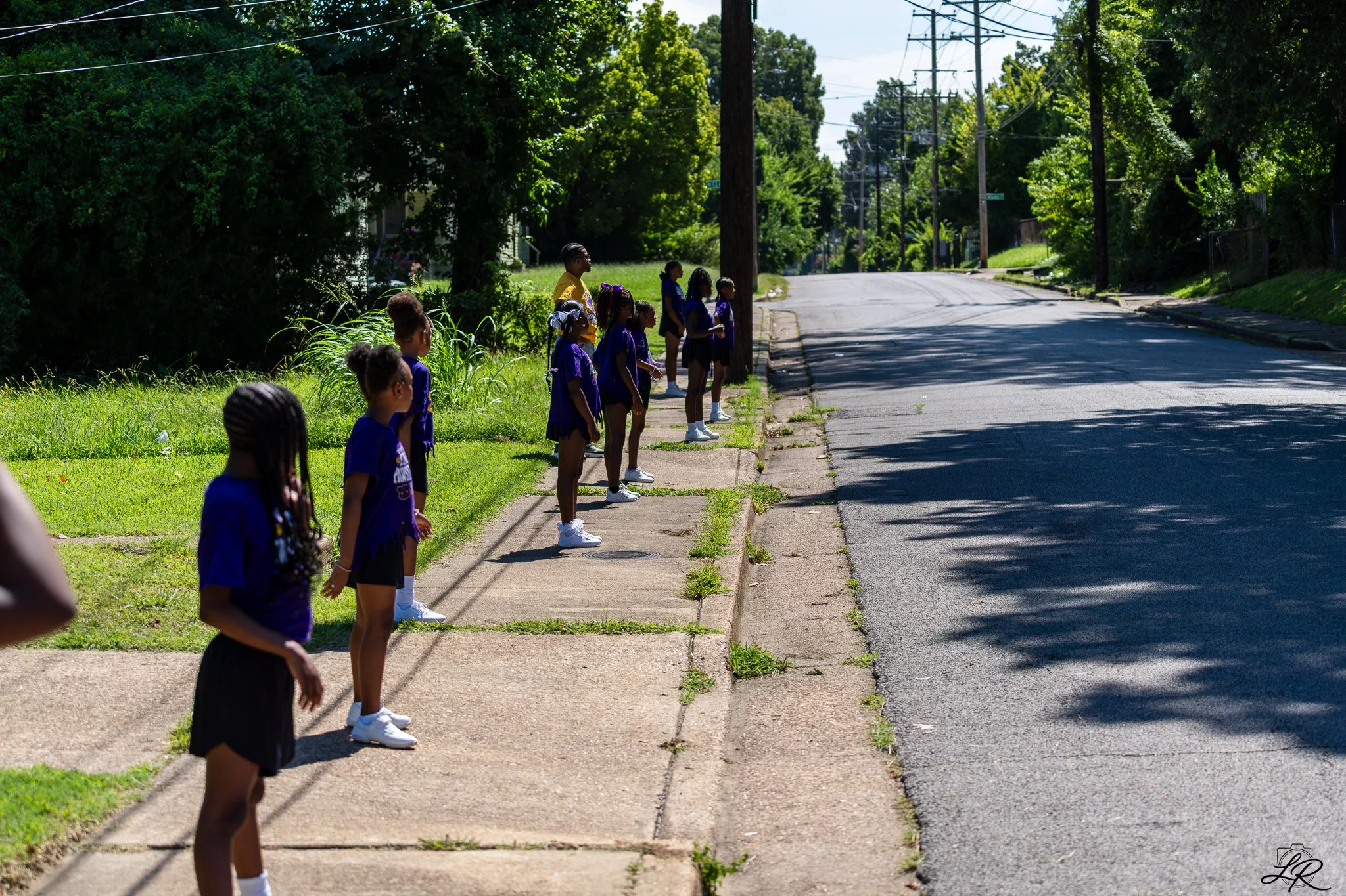 A group of young girls dressed in purple and black standing on a sidewalk next to a grassy area, waiting to cross the street on a sunny day.
