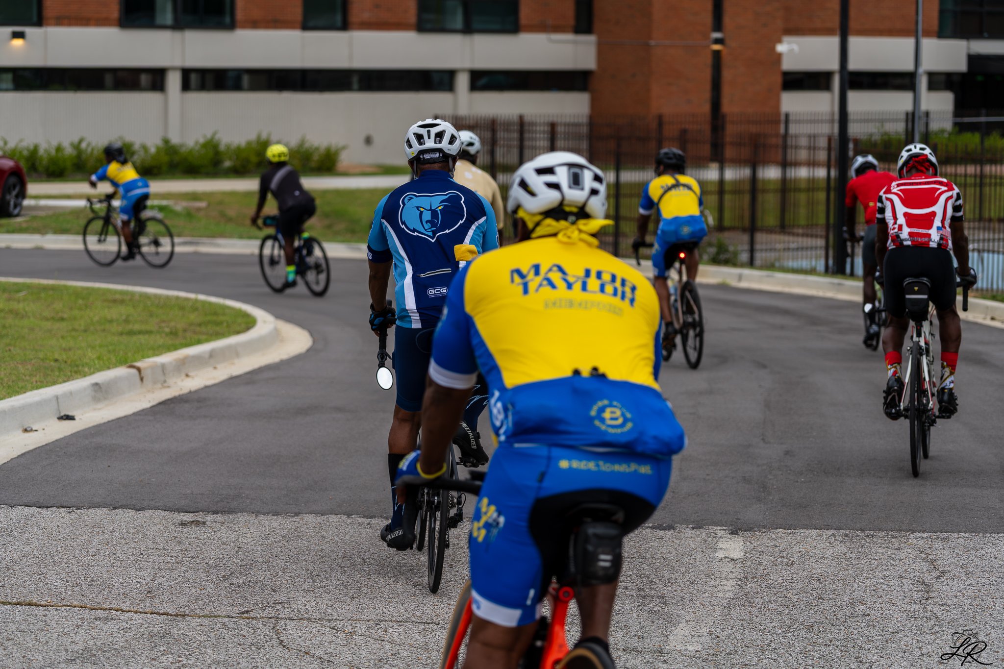 Group of cyclists riding on a paved road in an urban area, some wearing helmets and colorful jerseys, with buildings in the background.