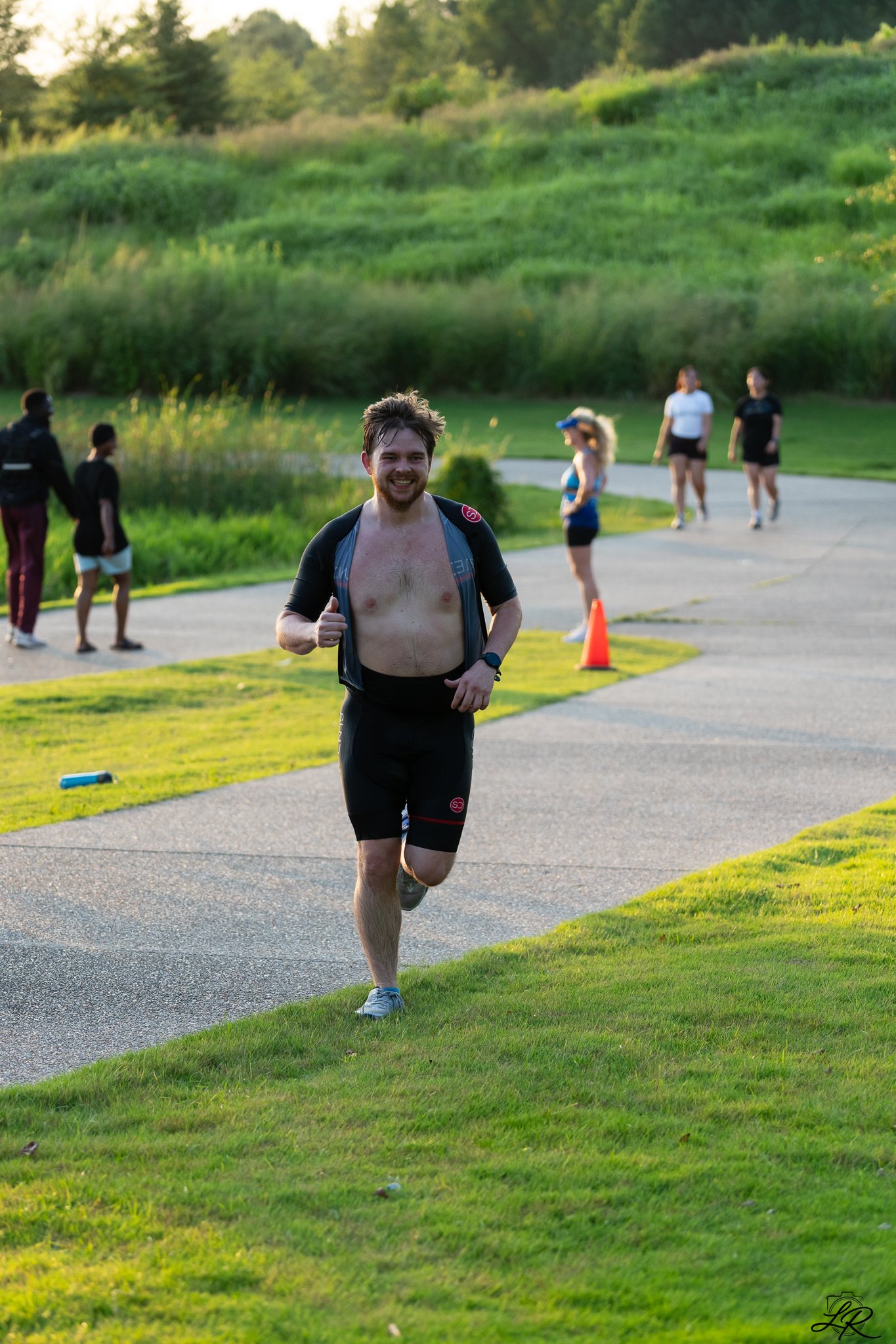 A man running outdoors during a race or event, smiling and shirtless, with others walking in the background on a paved path with green grass and trees.