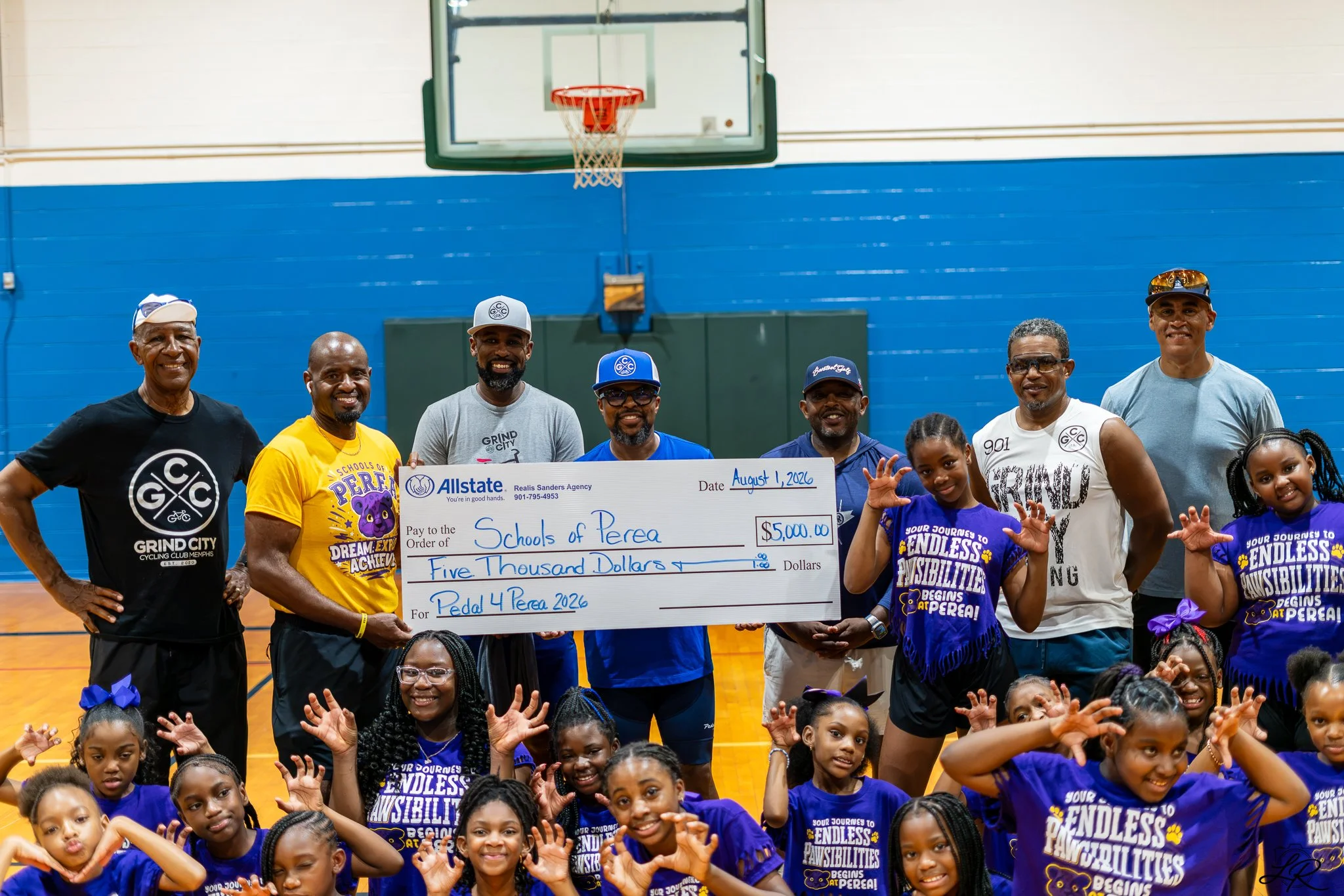A group of children and adults posing with an oversized check for five thousand dollars, awarded to Schools of Perea, in a basketball gymnasium. The children are wearing purple T-shirts with motivational slogans, and the adults are smiling and standi