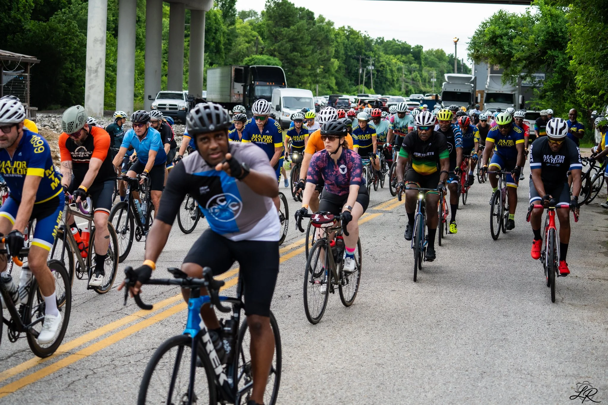 A large group of cyclists participating in a race or event on a paved road, wearing helmets and colorful athletic clothing, with trucks and green trees in the background under an overpass.