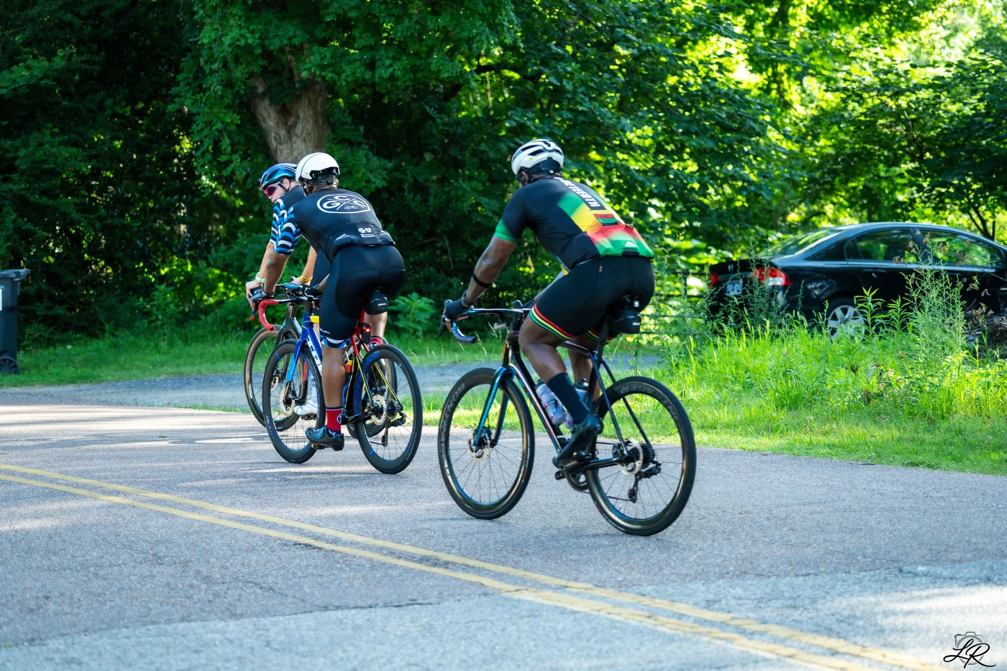 Three cyclists riding on a paved road surrounded by lush green trees during daytime. They are wearing helmets and athletic clothing.