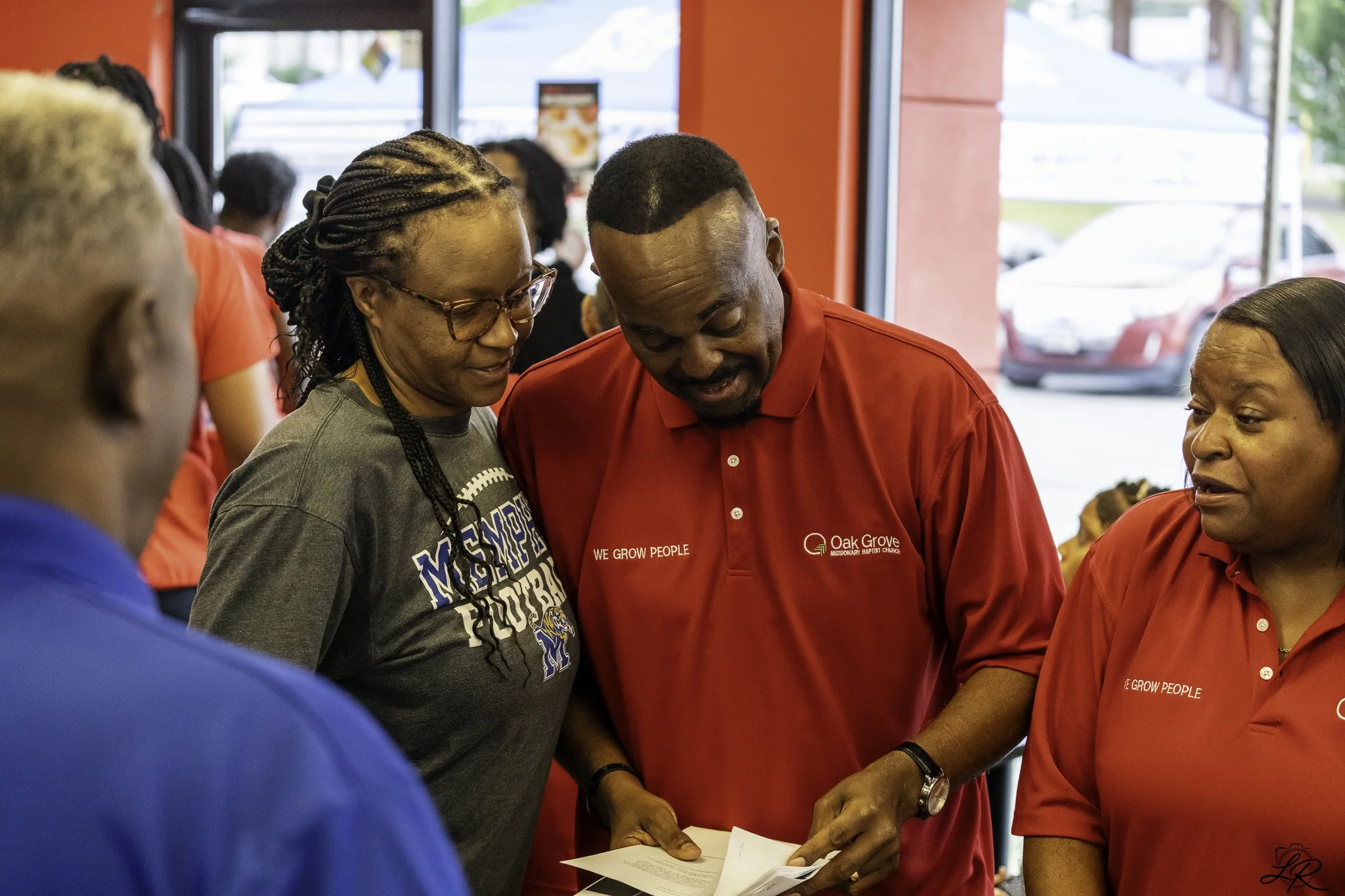 A group of people gathered inside a Red restaurant, with some wearing red shirts that have the words 'WE GROW PEOPLE' and 'Oak Grove Missionary Baptist Church' printed on them. They appear to be engaging in a discussion or sharing something.