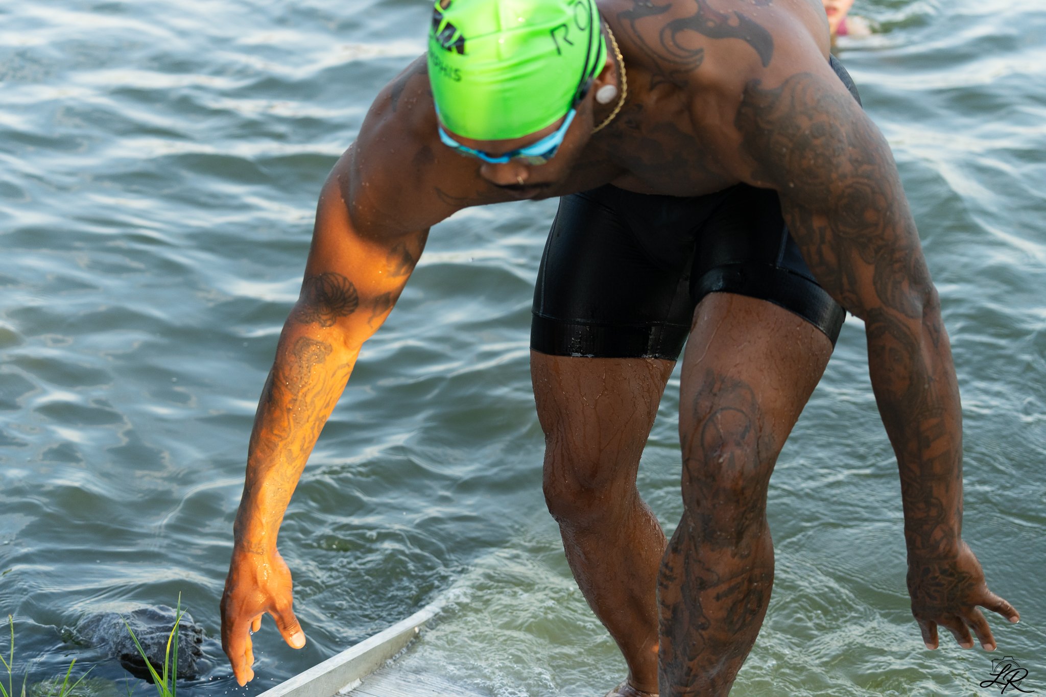 A swimmer wearing a bright green swim cap and goggles enters the water, showing their muscular build and numerous tattoos on their arms and torso.