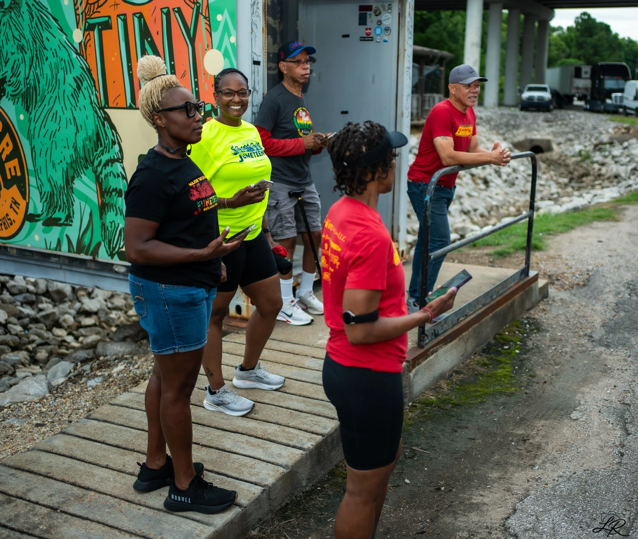 A group of five diverse people gathered outdoors on a wooden walkway, with a colorful mural in the background featuring a green bear and the words 'JUNETEEN.' They are wearing casual clothing, some with T-shirts supporting Juneteenth, and are looking