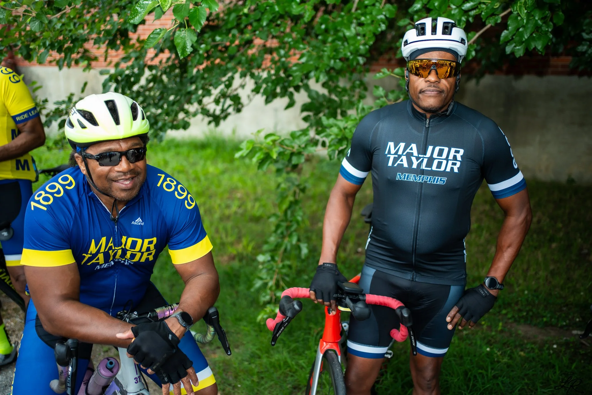 Two men wearing cycling helmets and sunglasses standing with bicycles outdoors near green bushes and a brick wall. One man is smiling, wearing a blue and yellow cycling jersey, and the other man has a serious expression, wearing a black cycling jerse