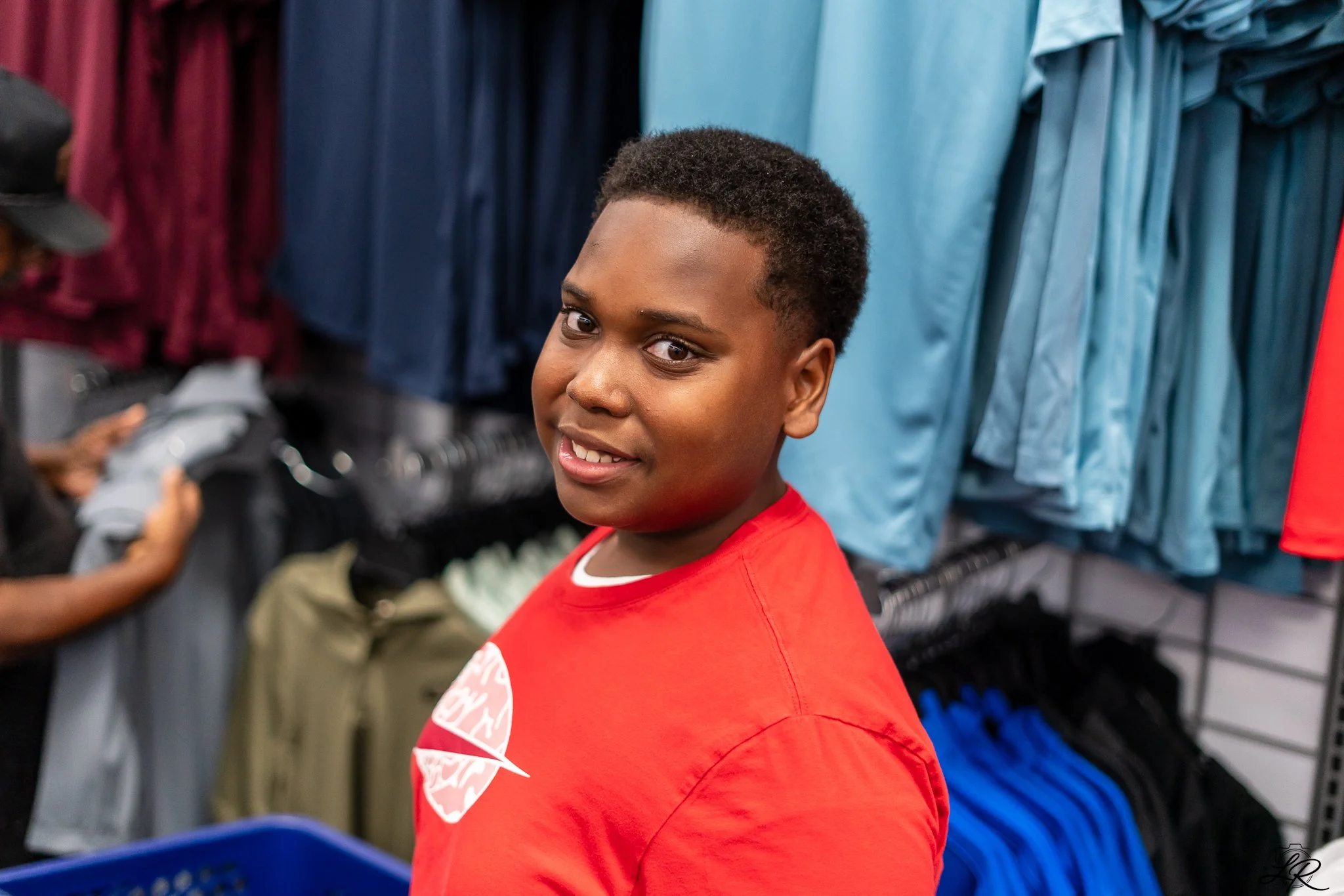 Young boy with short curly hair wearing a red shirt, smiling slightly, inside a clothing store with blue and maroon hanging clothes in the background.