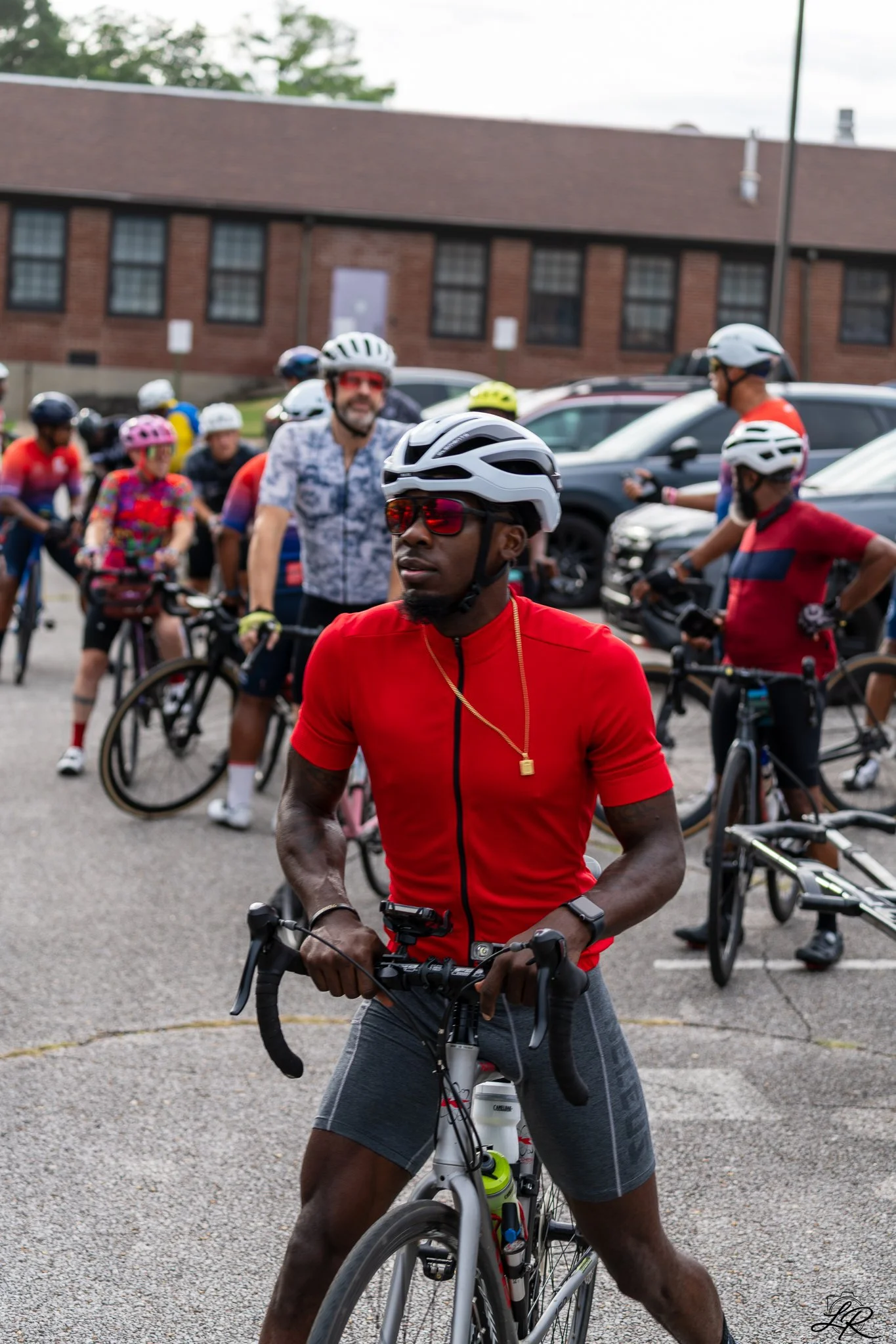 A group of cyclists gathered outdoors in a parking lot, preparing for a ride, with a brick building in the background.
