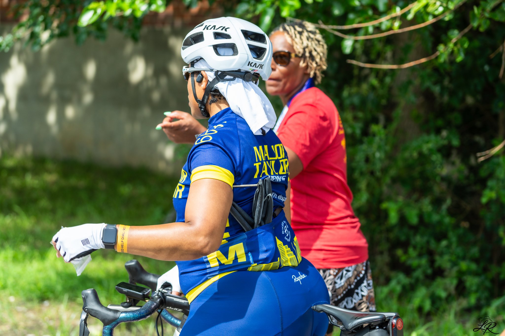 A woman in a blue cycling jersey and white gloves standing next to an electric bicycle, with a person in a red shirt and patterned skirt in the background, outdoors with green foliage.