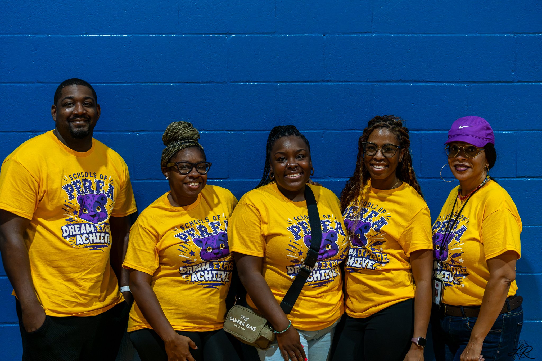 Six women and one man standing in front of a blue brick wall, all wearing yellow T-shirts with a purple panther graphic and text reading 'Schools of P.E.R.F.E.C.T. Dream, Explore, Achieve'.