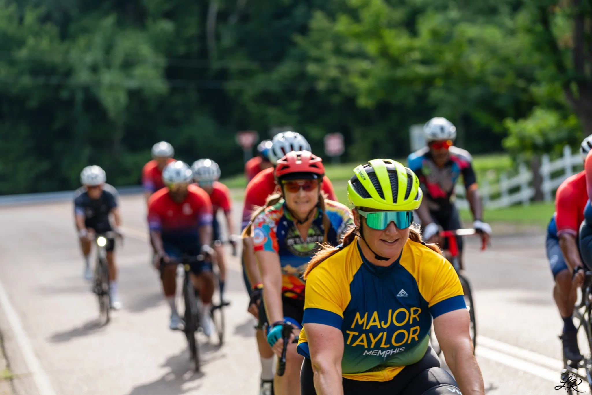 A group of cyclists riding on a road surrounded by green trees, with a woman wearing a yellow and blue cycling jersey in the foreground.