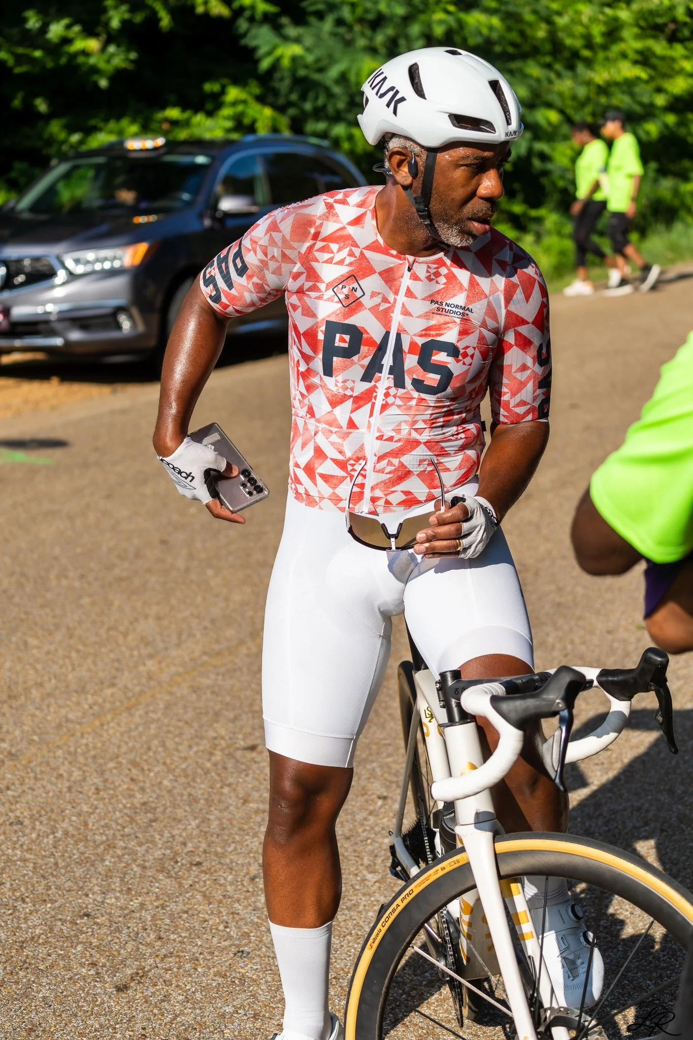 A man wearing a white helmet and cycling clothes stands with a bicycle outdoors, with two people in neon shirts walking and a black car in the background.