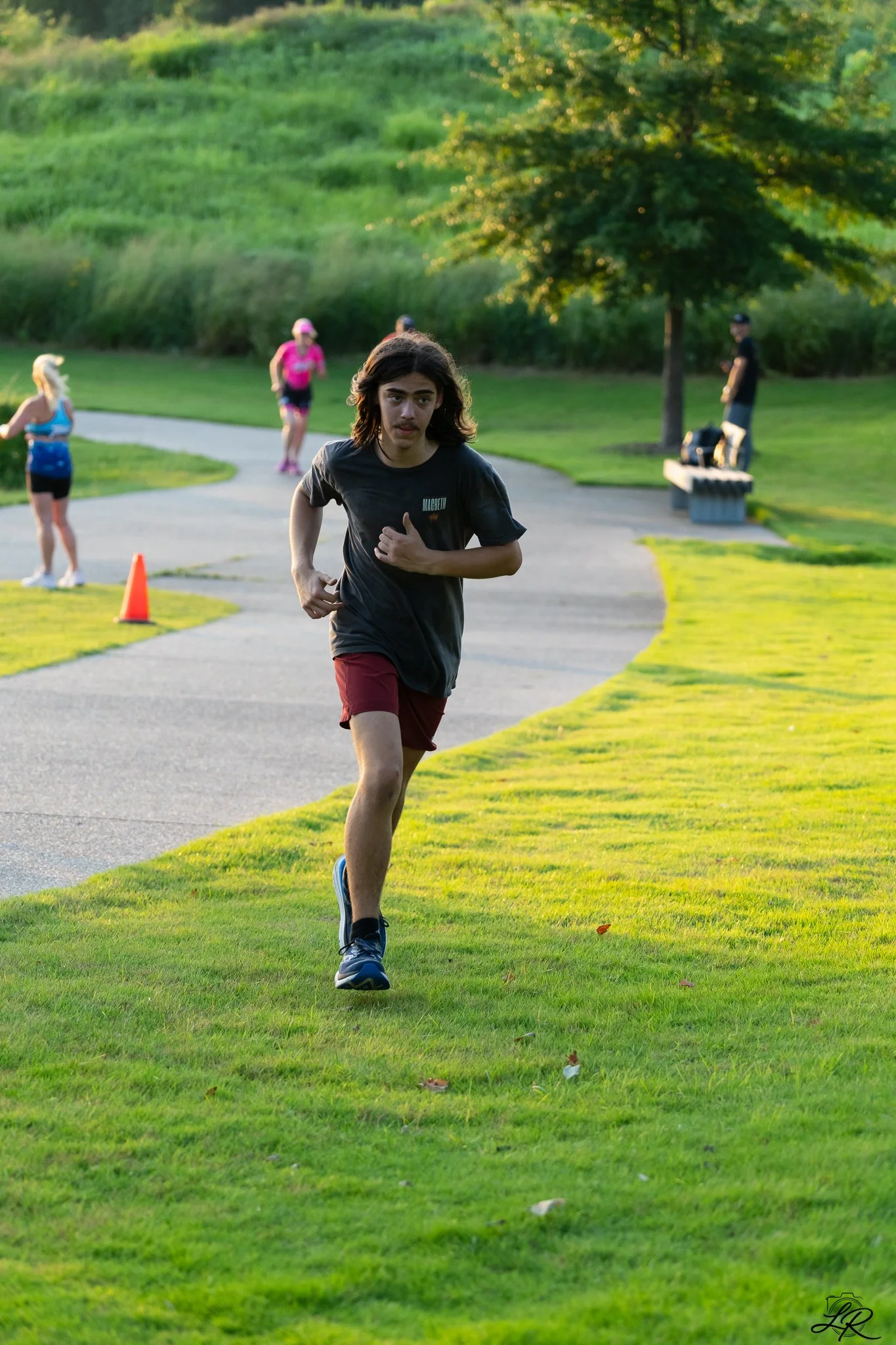 A young man running on a grassy park trail during sunset, with other people jogging or walking in the background, surrounded by trees and greenery.