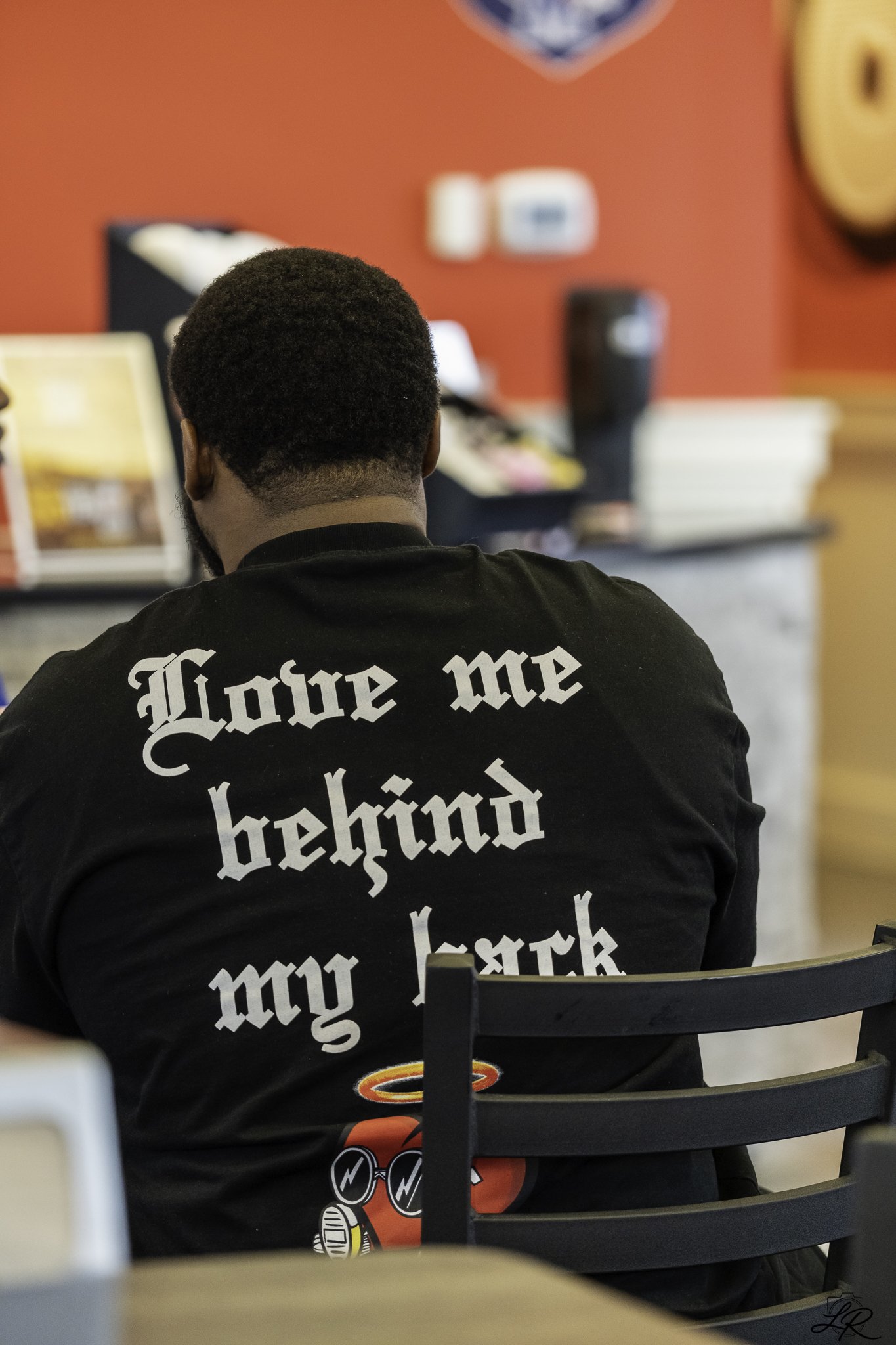 Back of a person wearing a black t-shirt with the phrase 'Love me better' in a gothic font, sitting on a black chair in front of a red wall with blurred objects and books.