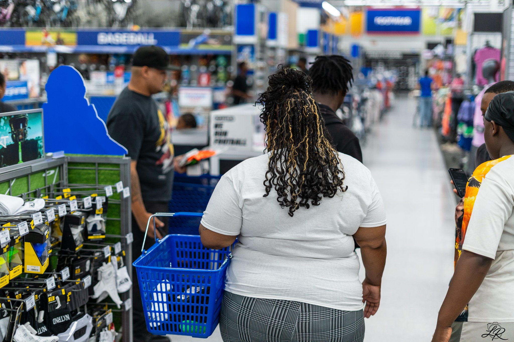 People shopping at a retail store, with a woman in the foreground carrying a blue shopping basket and a man beside her using his phone, surrounded by shelves of products.