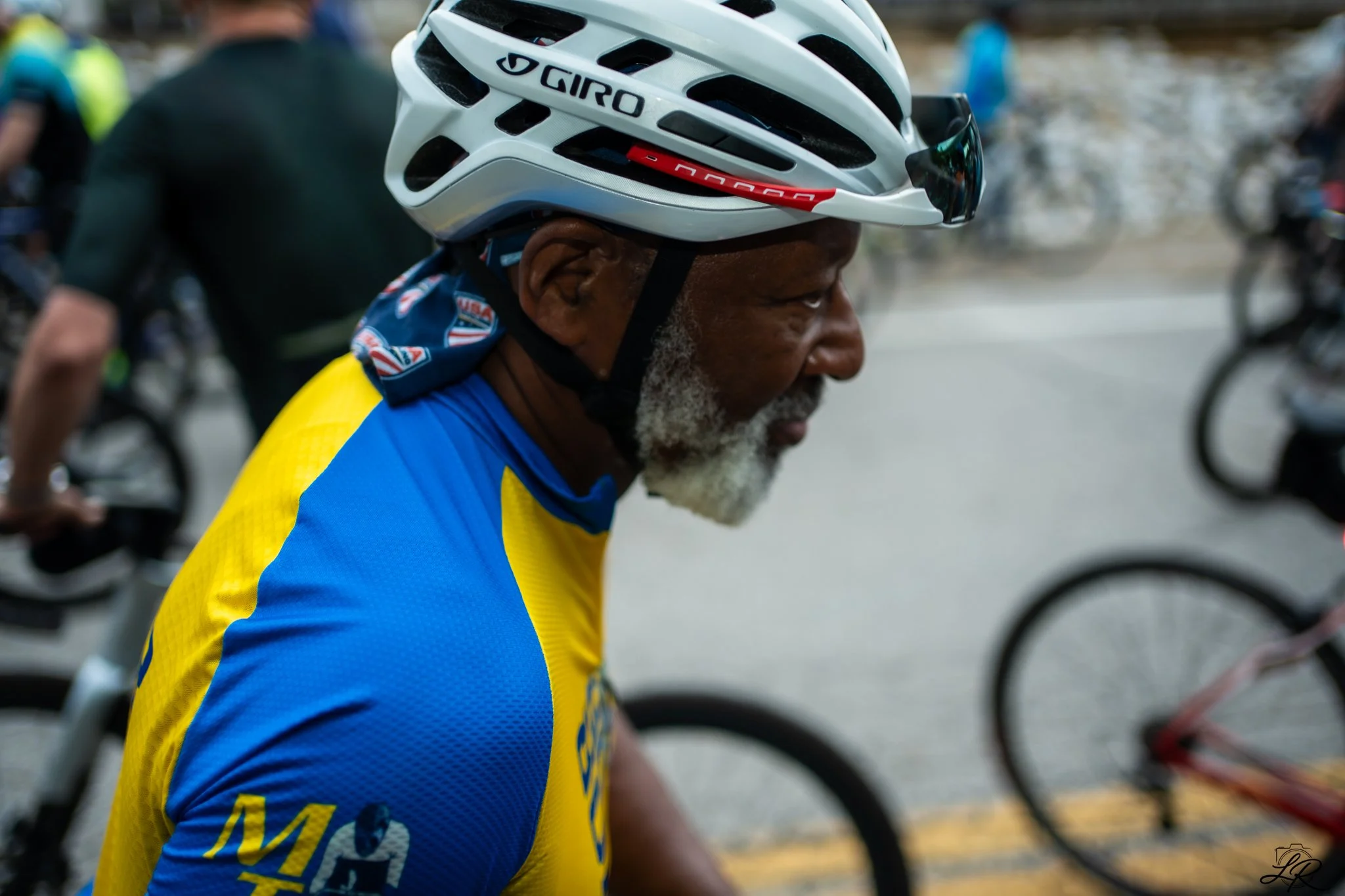 A side profile of an elderly man wearing a white cycling helmet and a blue and yellow cycling jersey, surrounded by other cyclists during a biking event.