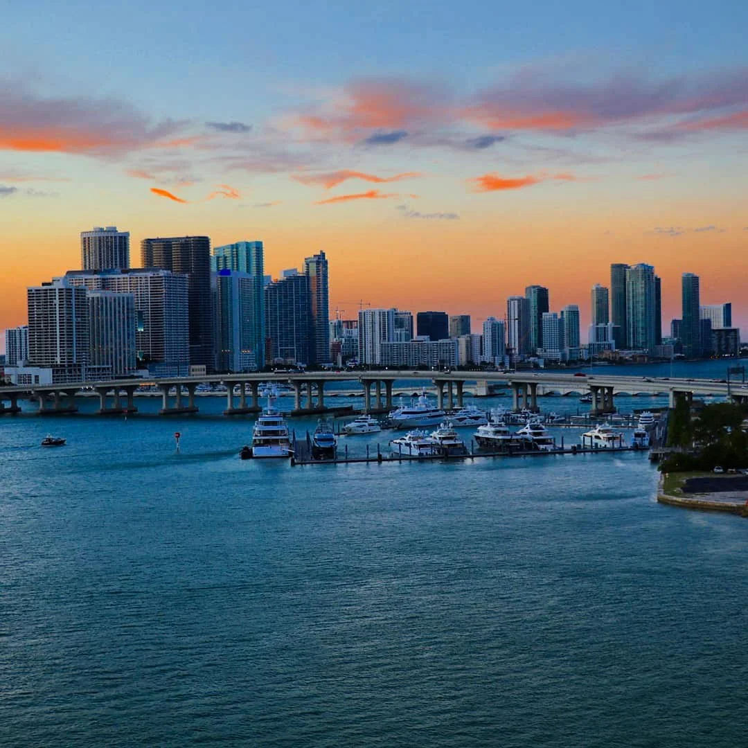A city skyline during sunset with high-rise buildings, a bridge, and a marina filled with yachts and boats on the water